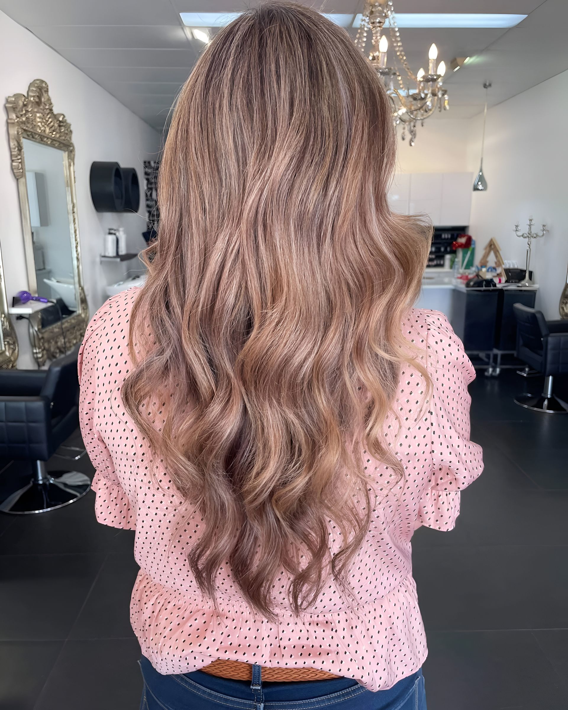 A person with long, crimped, light brown wavy hair, wearing a pink patterned top, standing inside a hair salon.