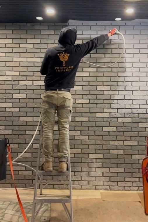 Une personne portant un sweat à capuche pulvérise du spray sur un mur de briques tout en se tenant sur un escabeau.
