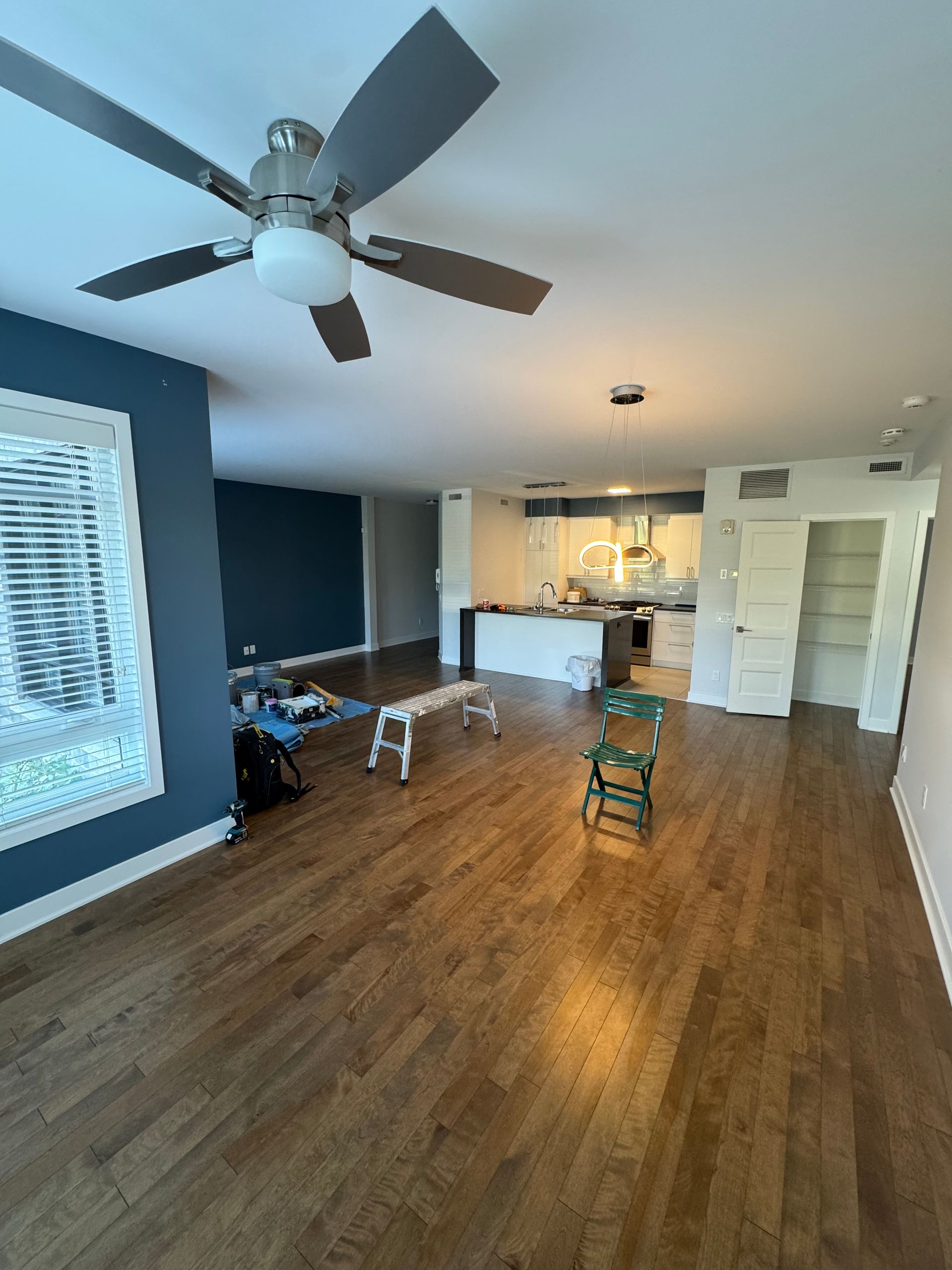 Espace de vie ouvert avec mur d'accent bleu, planchers de bois franc, ventilateur de plafond et cuisine en arrière-plan.