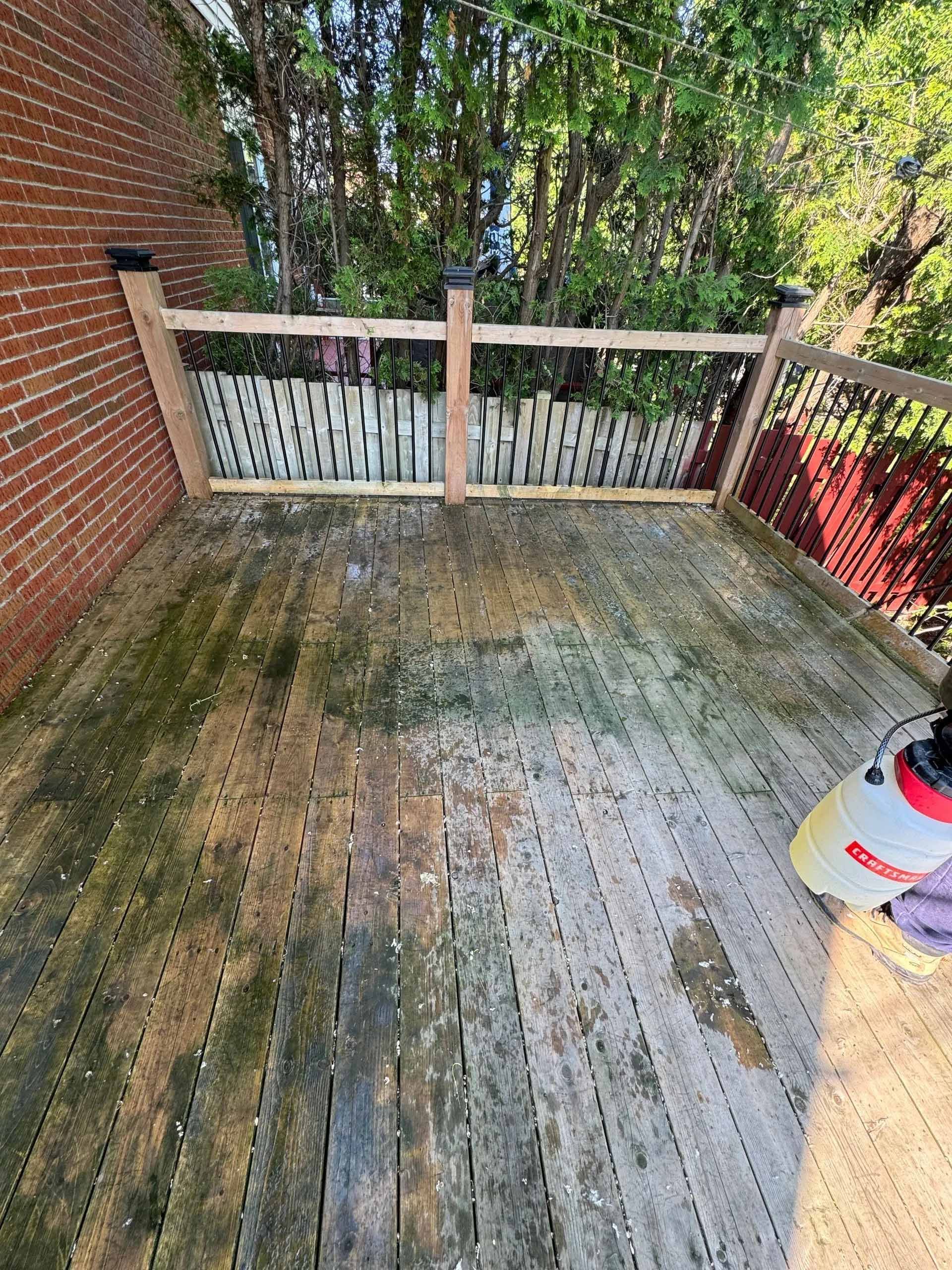 Une terrasse en bois, partiellement nettoyée, avec un pulvérisateur. Un mur de briques et une clôture sont visibles en arrière-plan.