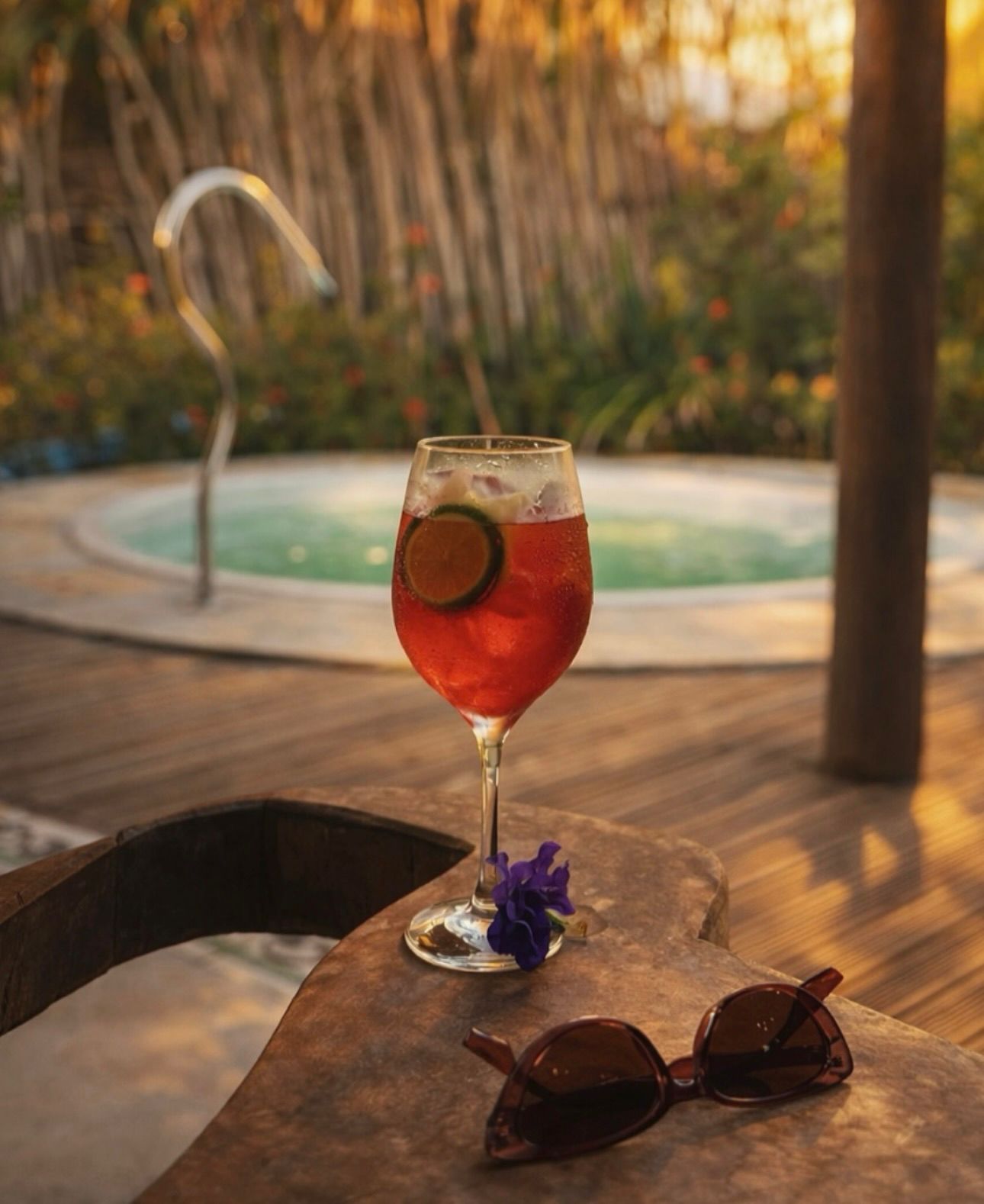 Um coquetel avermelhado com uma fatia de limão e uma flor roxa repousa sobre uma mesa de madeira ao lado de óculos de sol, com uma piscina ao fundo.