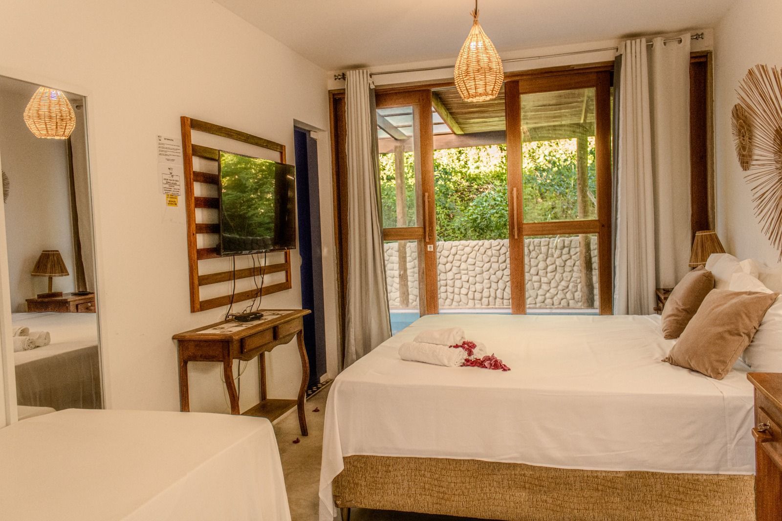 Um quarto de hotel com uma cama branca, uma pequena escrivaninha com televisão, um espelho e grandes portas de vidro que dão para um terraço.