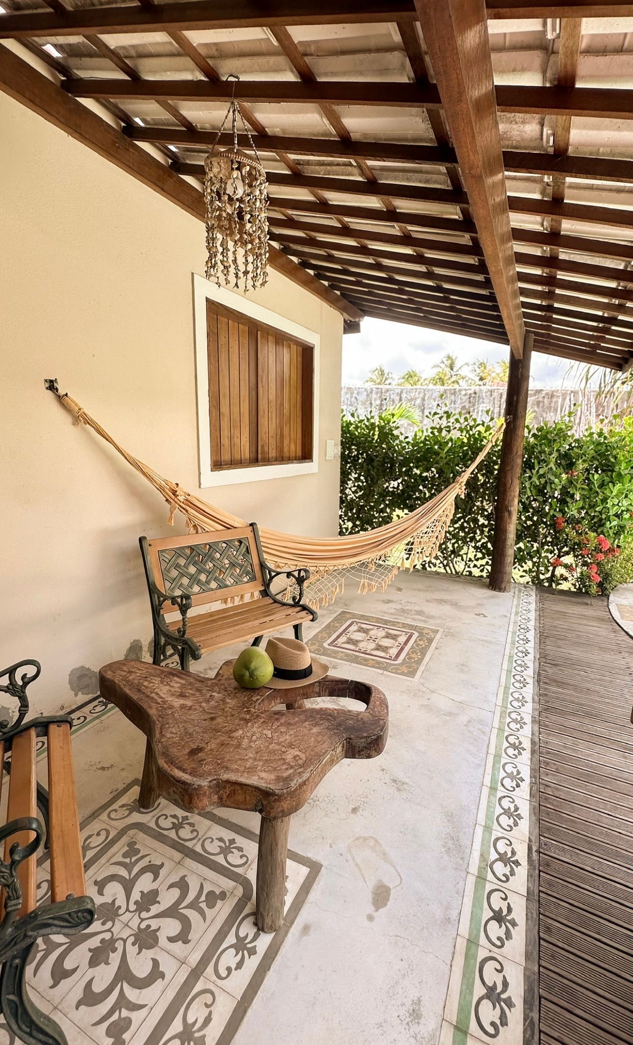 Uma varanda ensolarada conta com uma mesa de madeira, um banco, uma rede e piso de ladrilhos estampados sob uma pérgola rústica de madeira.
