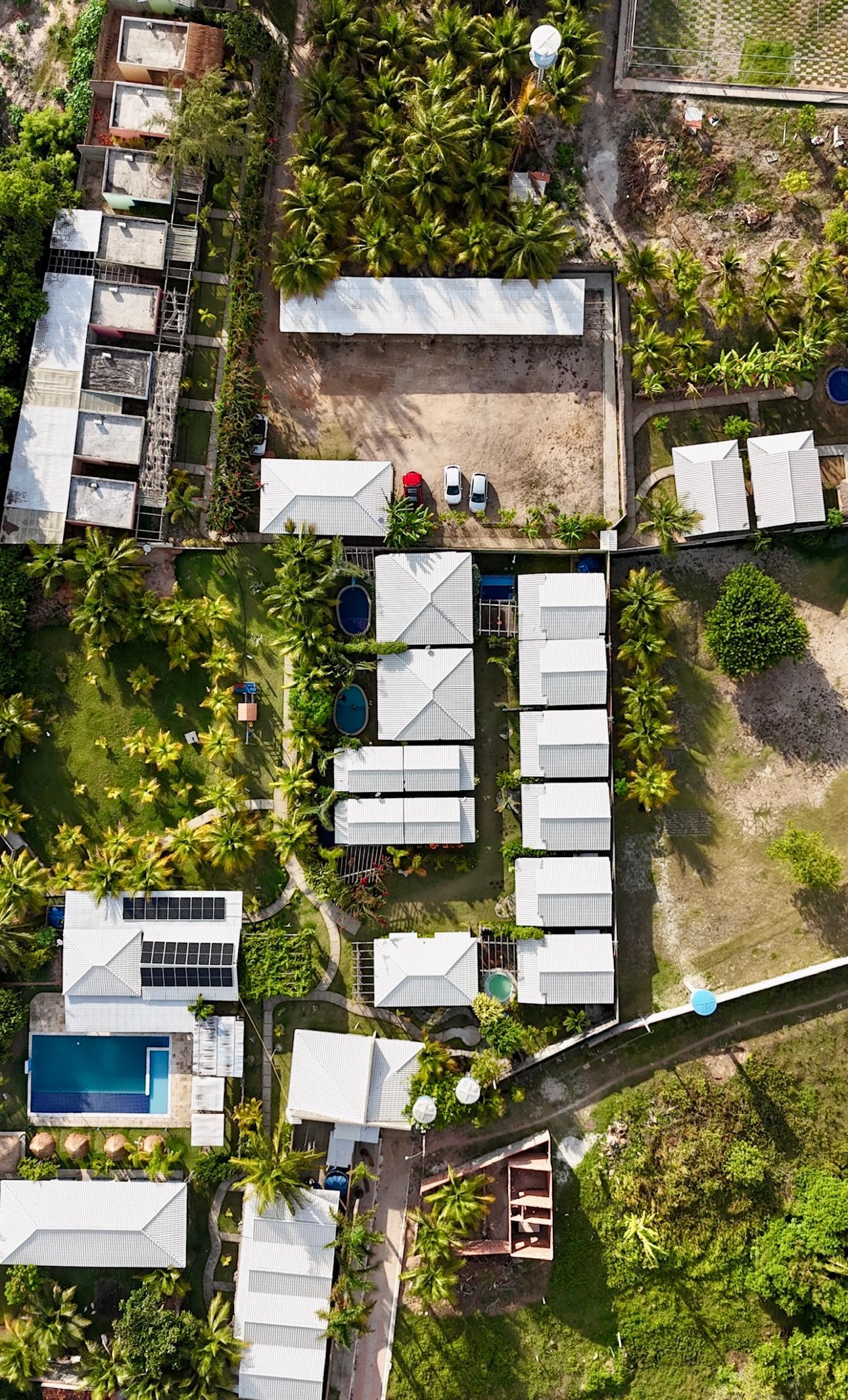 Vista aérea de uma propriedade exuberante com vários edifícios de telhados brancos, jardins verdejantes e uma piscina.
