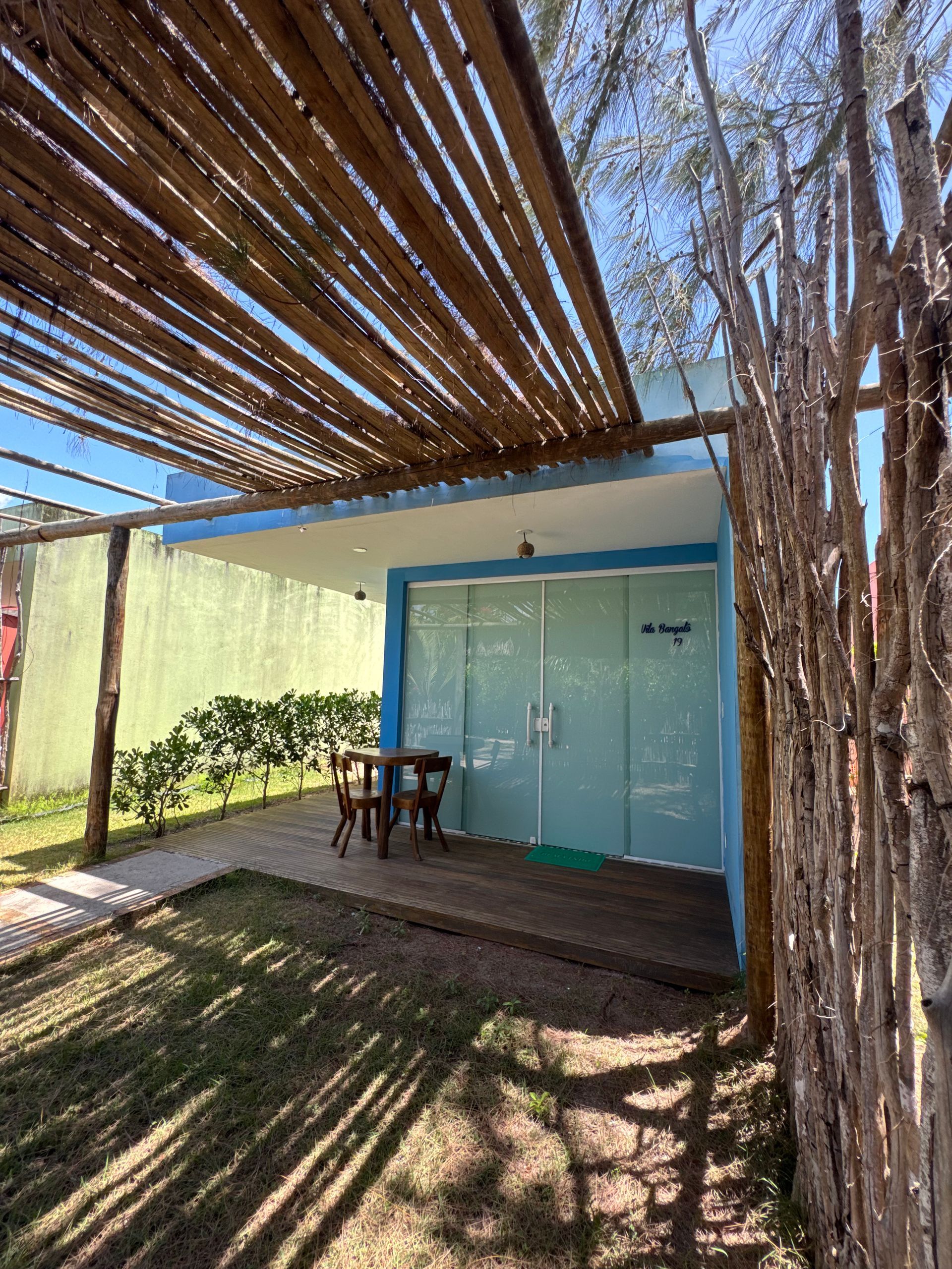 Uma pequena cabana azul com portas de vidro, um deck de madeira e uma pérgola de bambu, situada num quintal com uma cerca rústica de madeira.