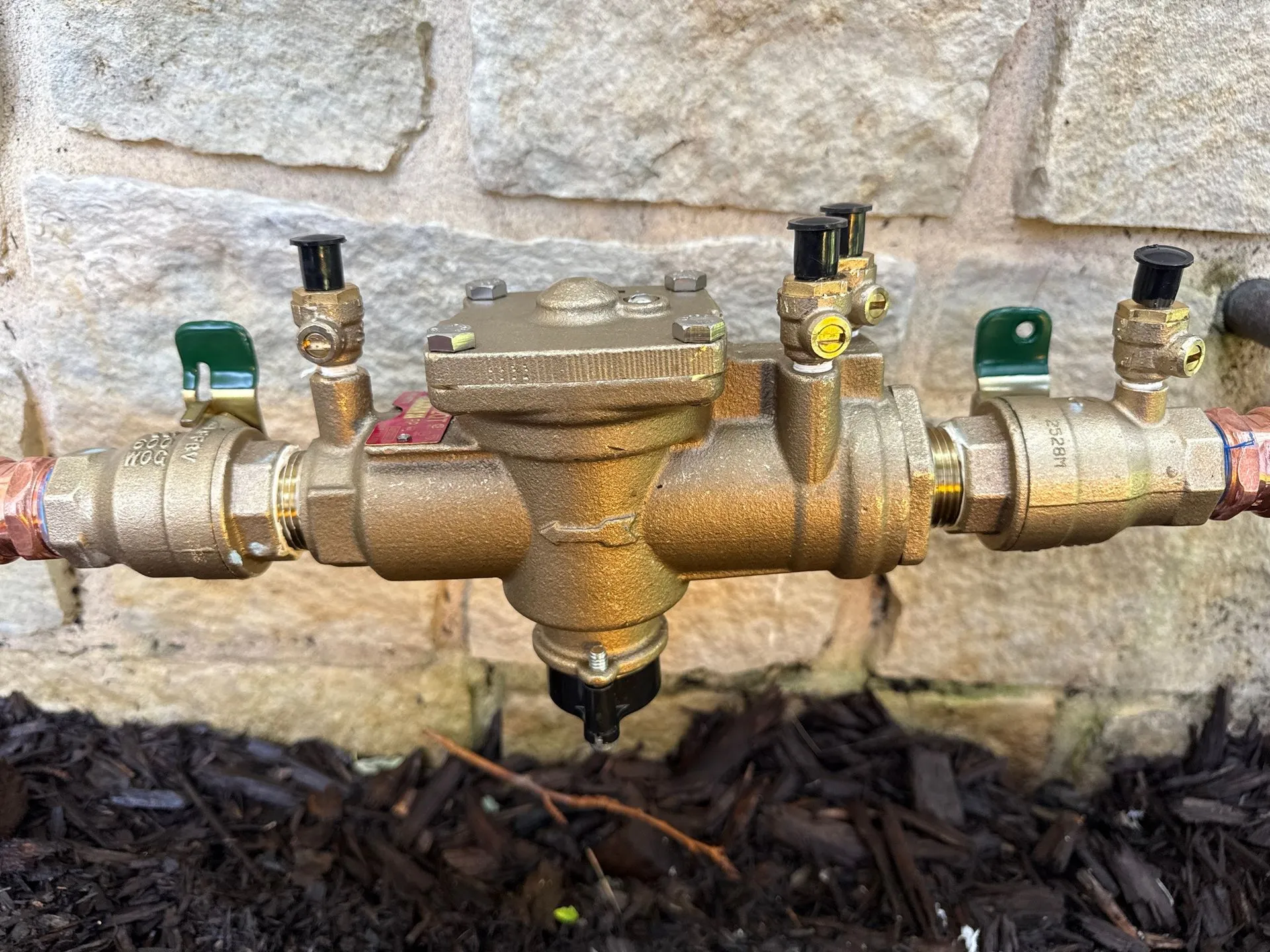 Bronze backflow preventer with valves and pipes against a stone wall.