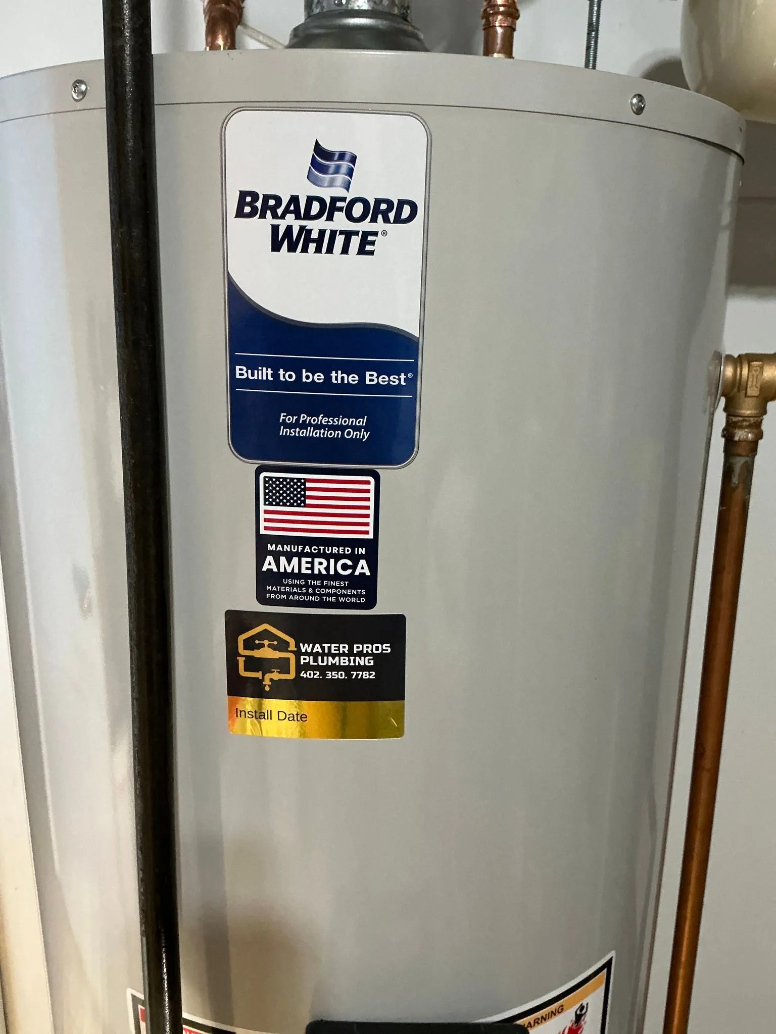 A Bradford White water heater in a utility room. Gray tank with brand decals. Copper pipes.