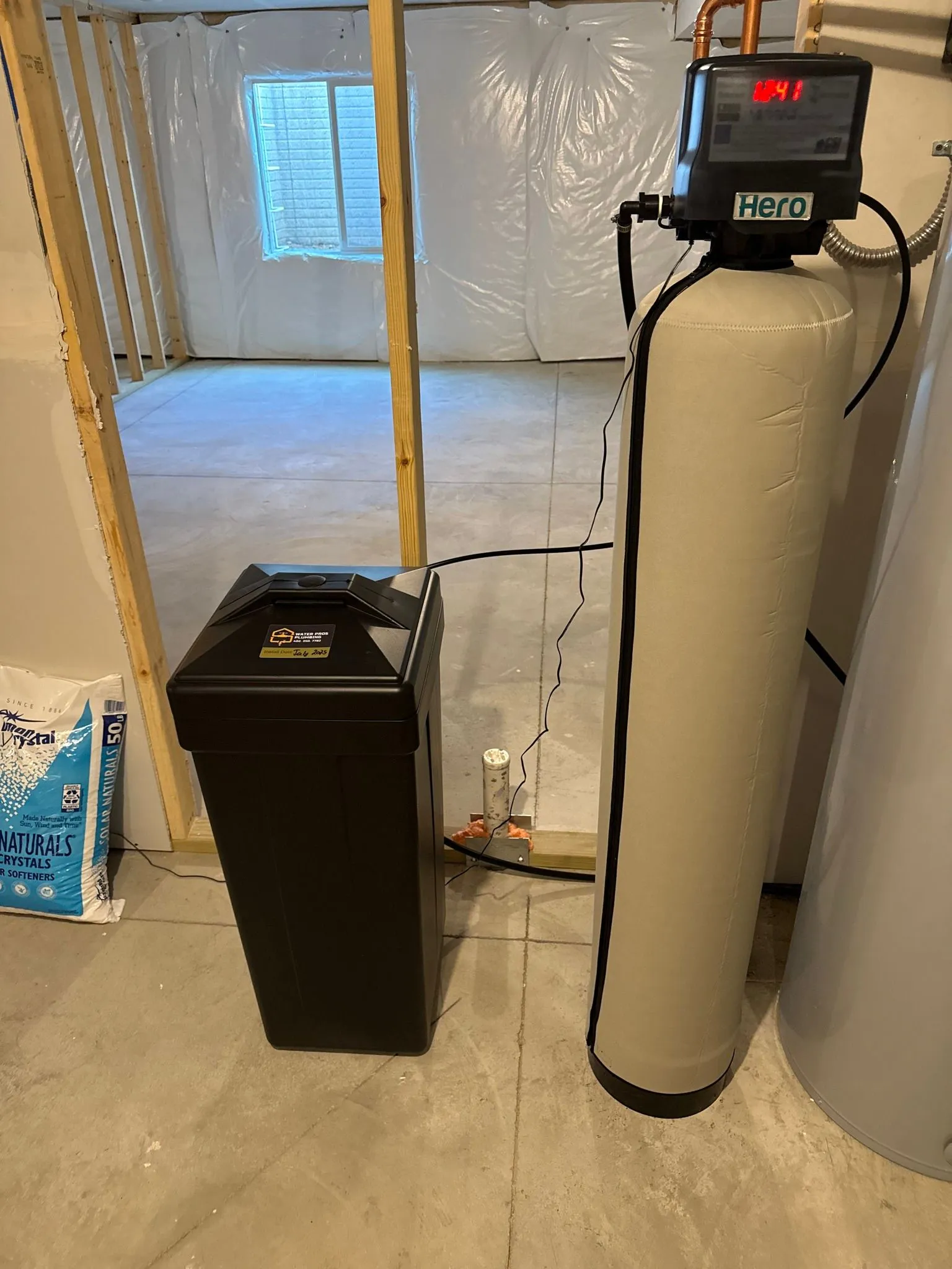 Water softener system in a basement setting with salt tank. Tall, beige tank and black control unit.