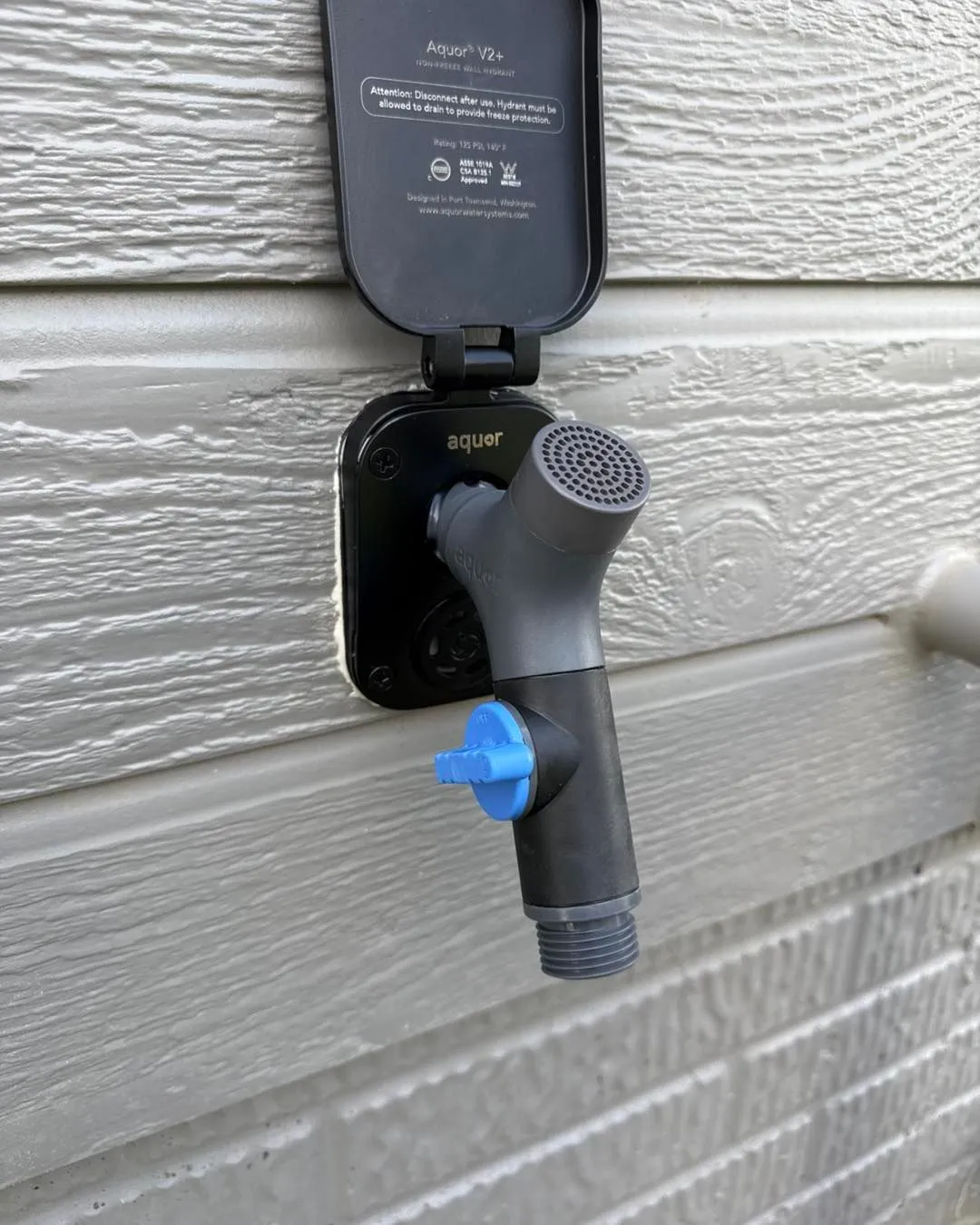 Outdoor faucet with a black cover and gray and blue components on a siding wall.
