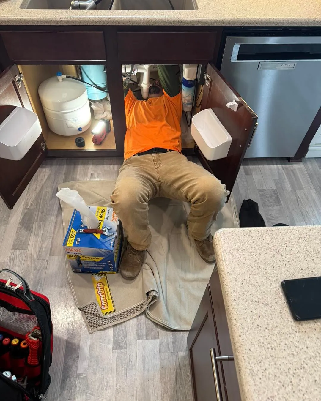 A person works under a kitchen sink, wearing an orange shirt and brown pants.