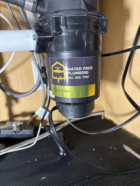 Black garbage disposal unit under a sink with plumbing and electrical wires.