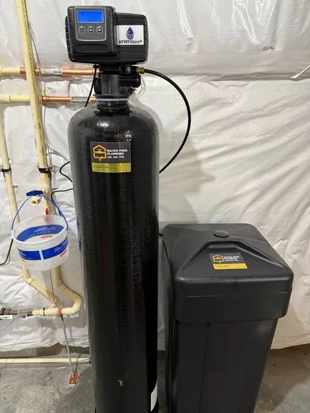 Water softener system with black tank, control panel, and salt storage bin. Pipes and a white wall are in the background.