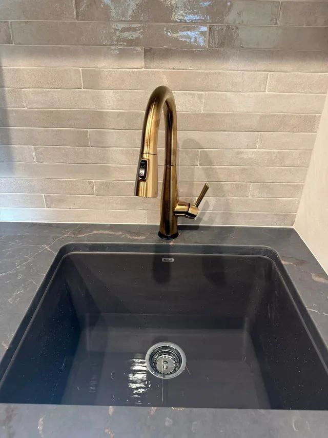 Gold kitchen faucet over a dark gray sink, set against gray tiled backsplash and dark gray countertop.
