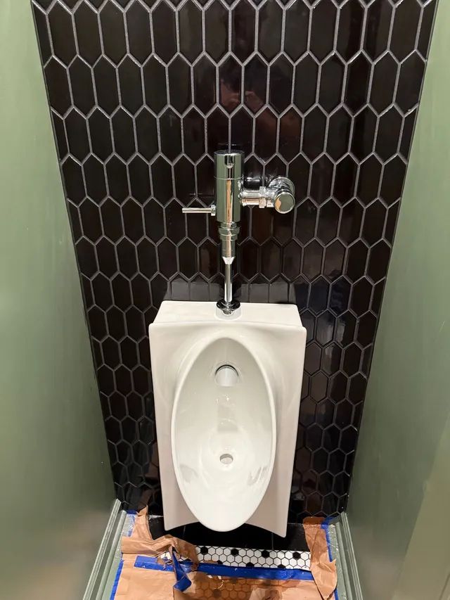 Urinal with black tile backdrop, mounted on a wall with plumbing and flush valve.