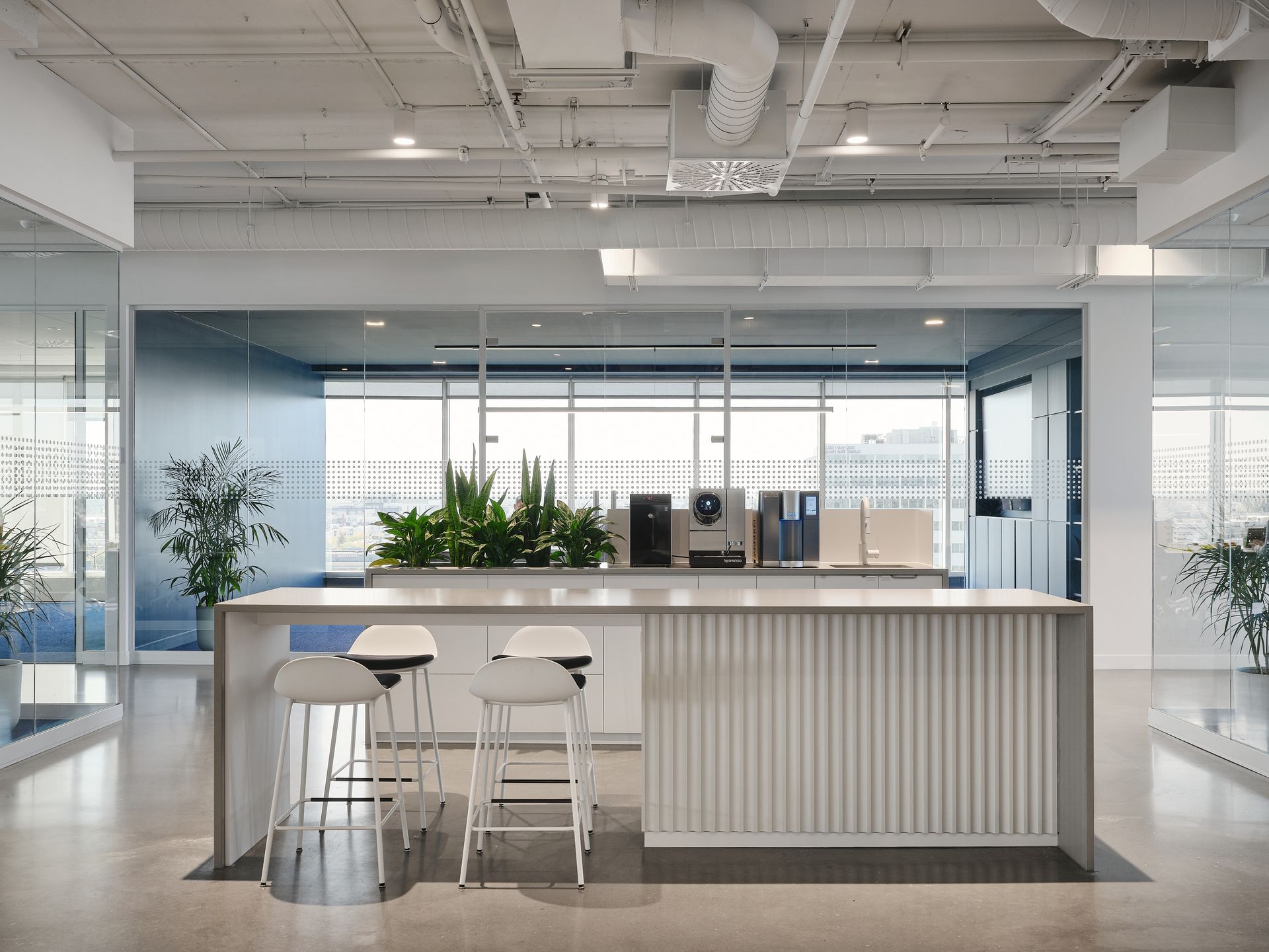 Espace détente moderne au bureau avec comptoir, tabourets, plantes et grandes fenêtres.