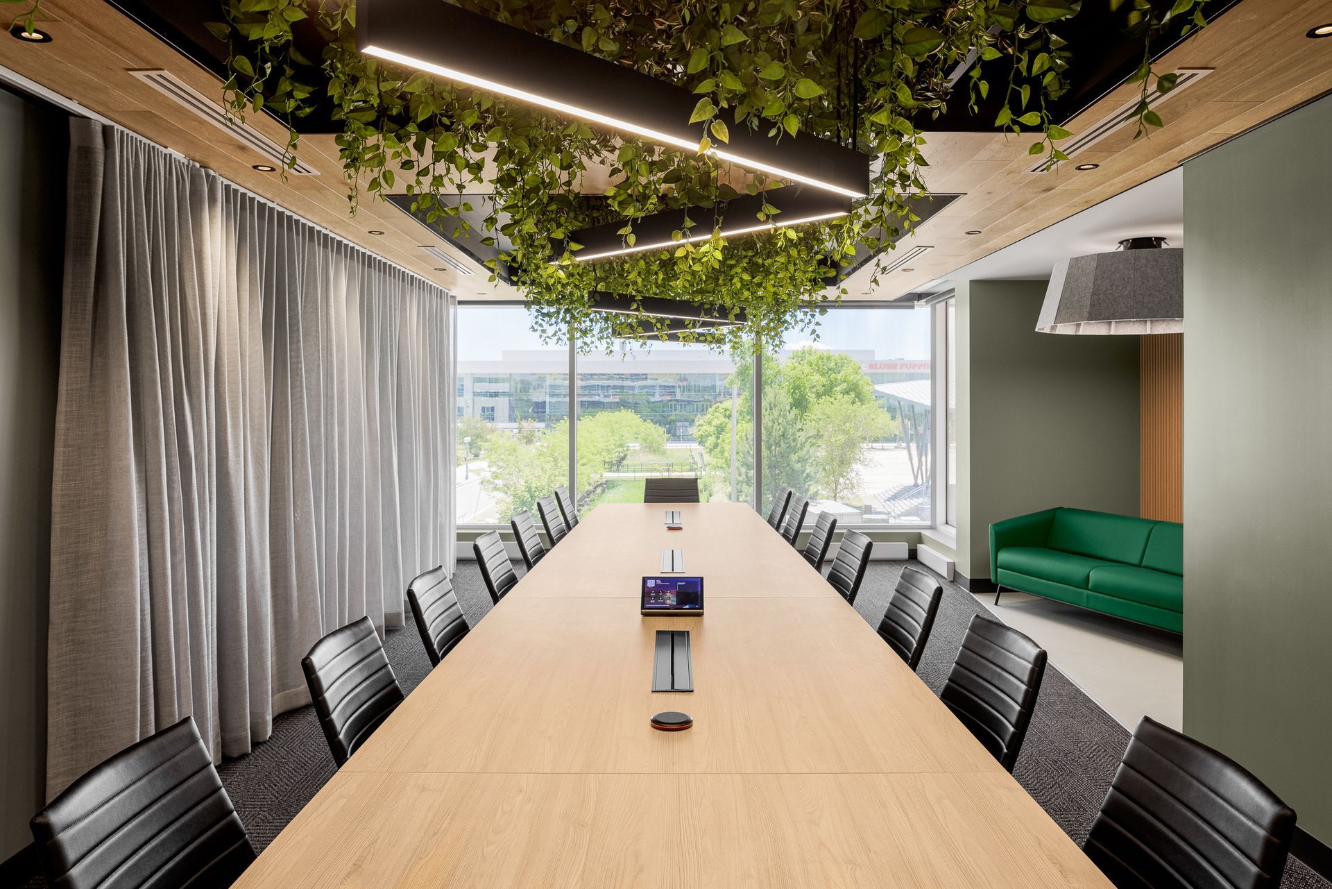 Salle de conférence avec une longue table, des chaises noires, un canapé vert, une grande fenêtre et des plantes suspendues au plafond.