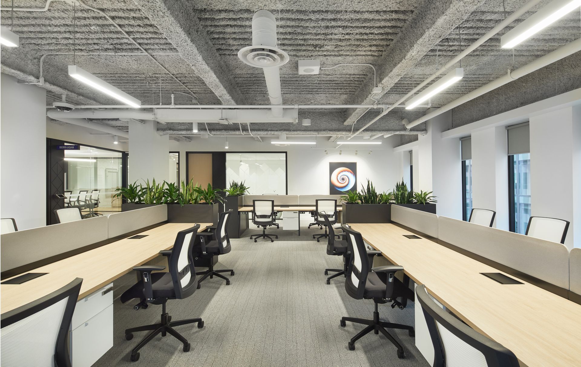 Espace de bureaux moderne avec de longs bureaux, des chaises noires, des plantes et un plafond apparent.