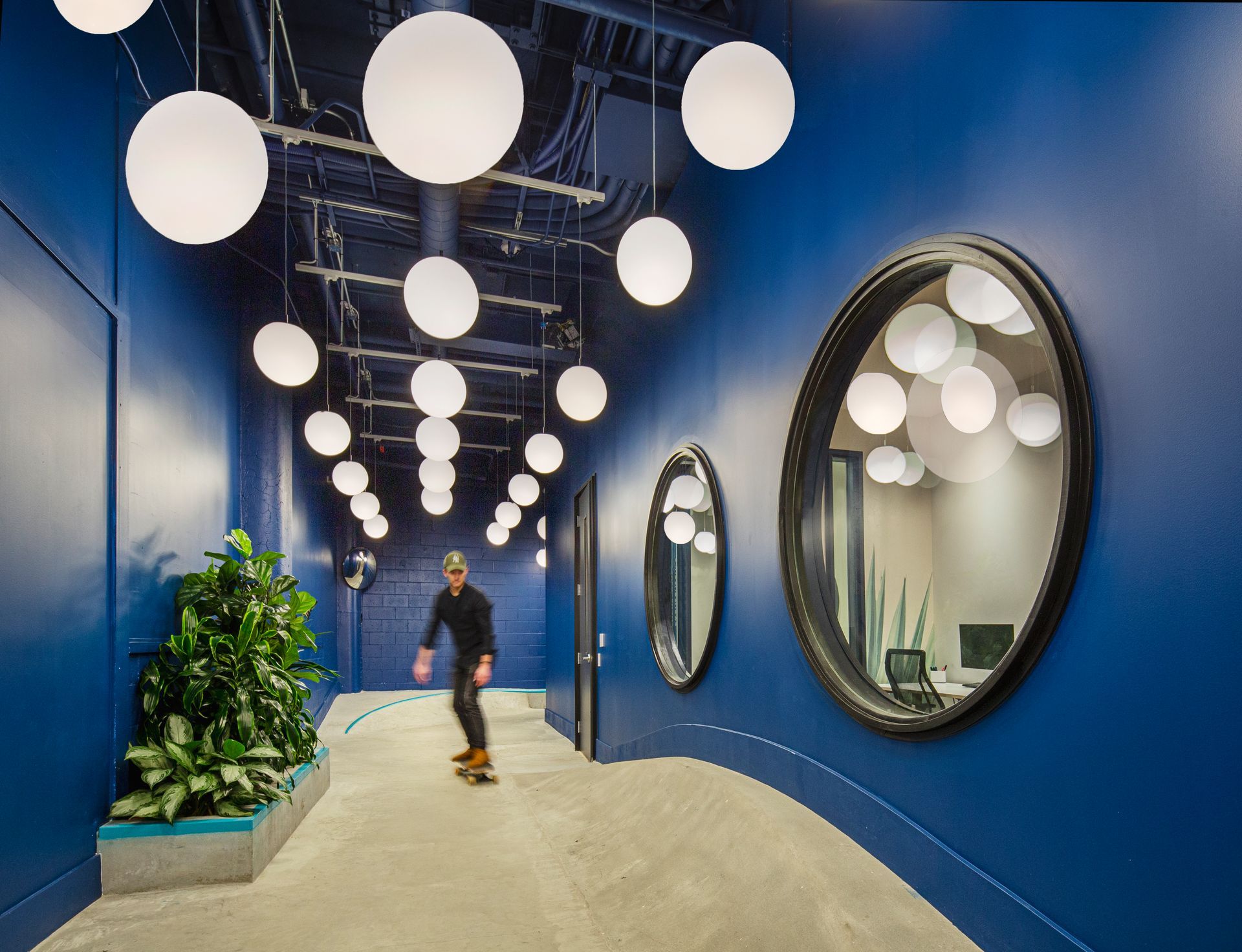 Couloir bleu avec luminaires ronds, miroirs et une personne faisant du skateboard. Plantes à gauche.