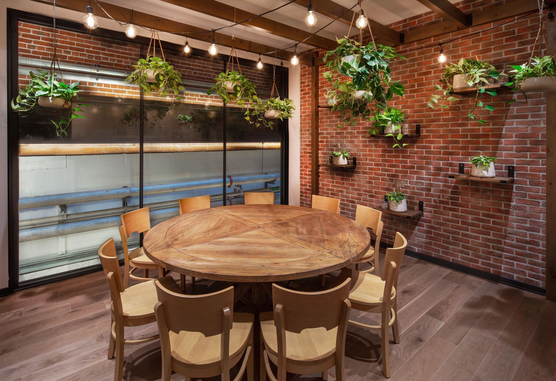 Une salle à manger avec une grande table en bois entourée de chaises, des murs en briques et des plantes suspendues.