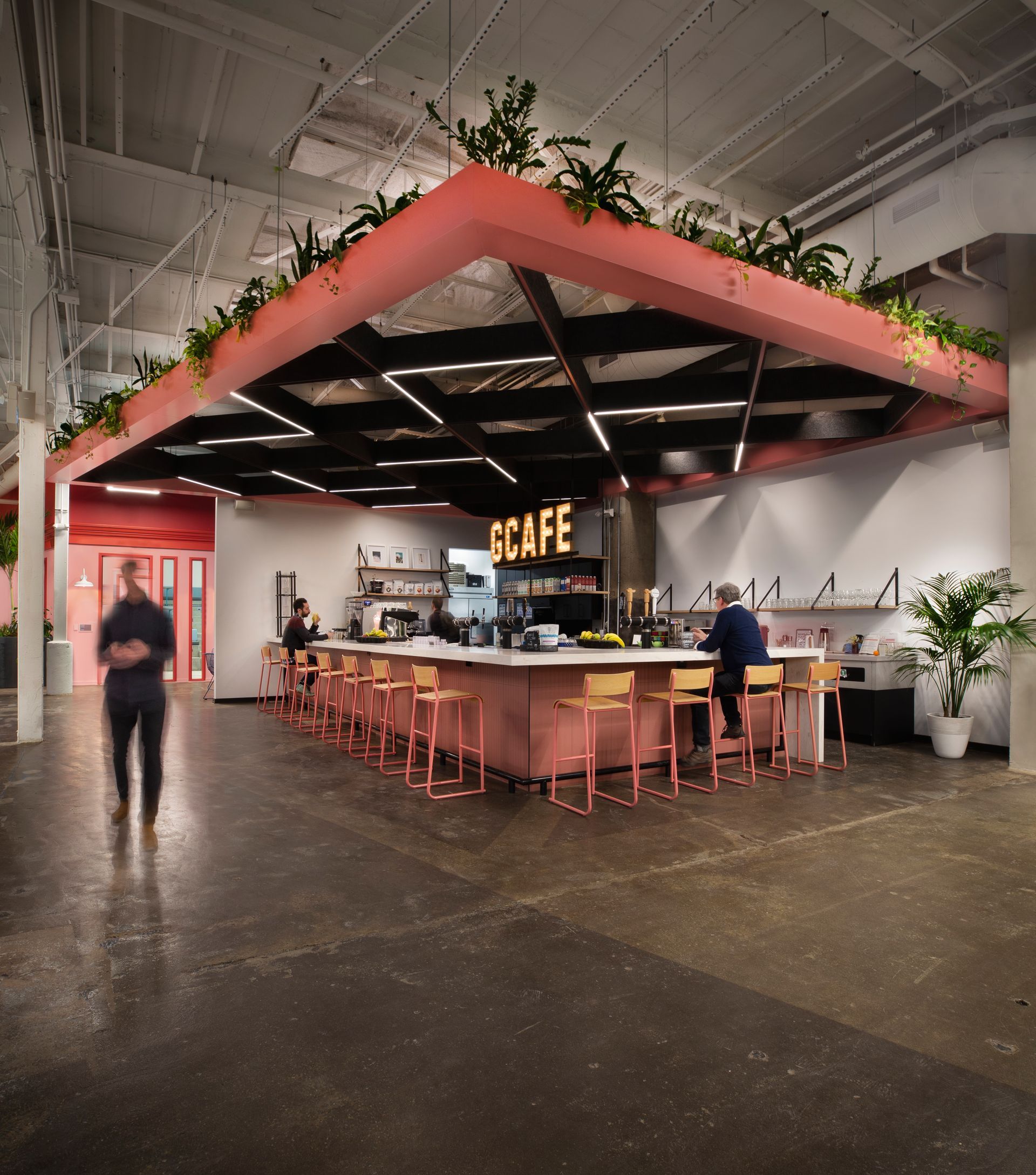Intérieur du café avec des touches de corail : bar, sièges et structure suspendue ornée de plantes. On aperçoit des clients.
