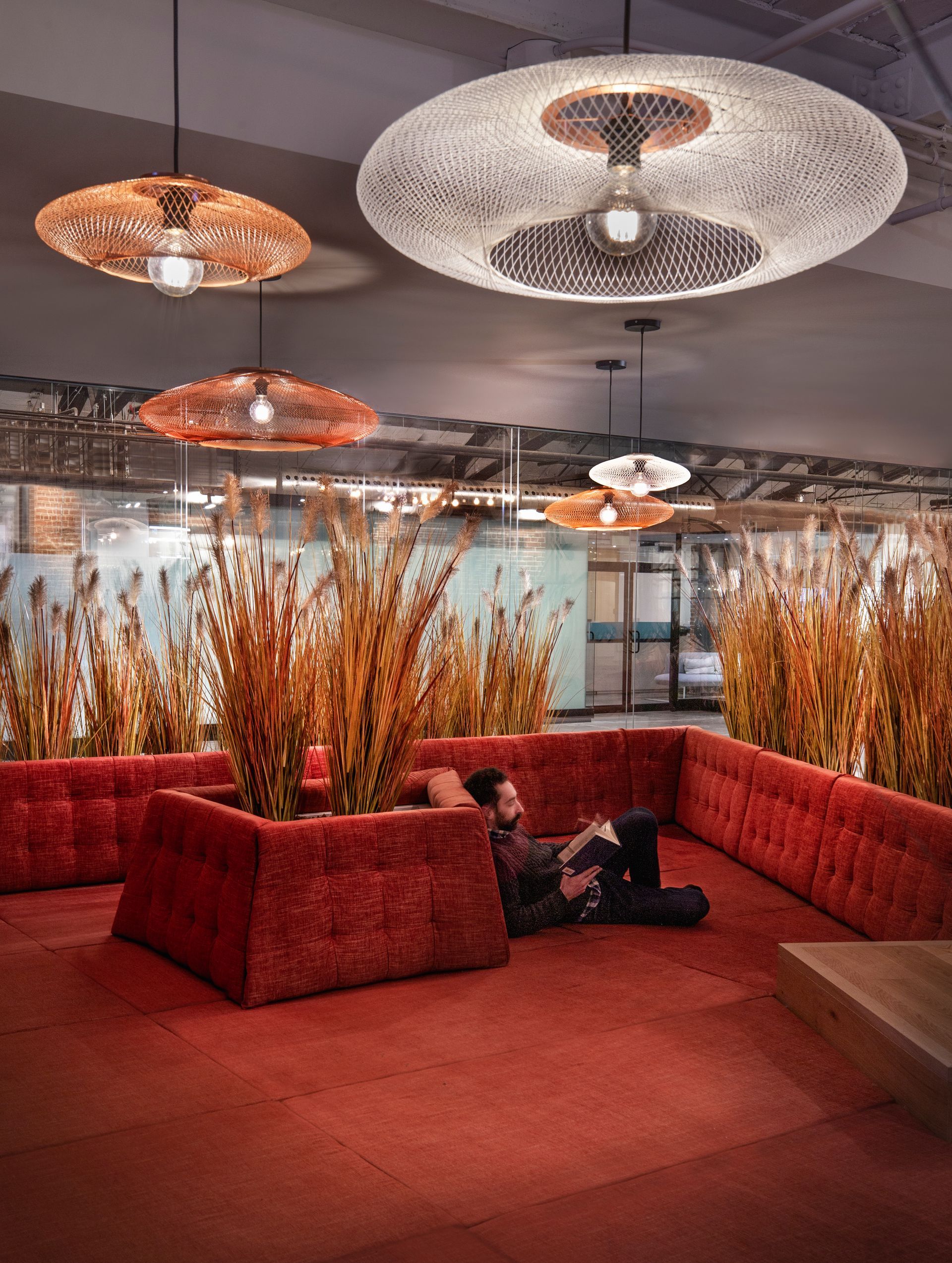 Une personne se détend sur un canapé rouge sous des suspensions lumineuses à motifs. De hautes herbes agrémentent le décor.