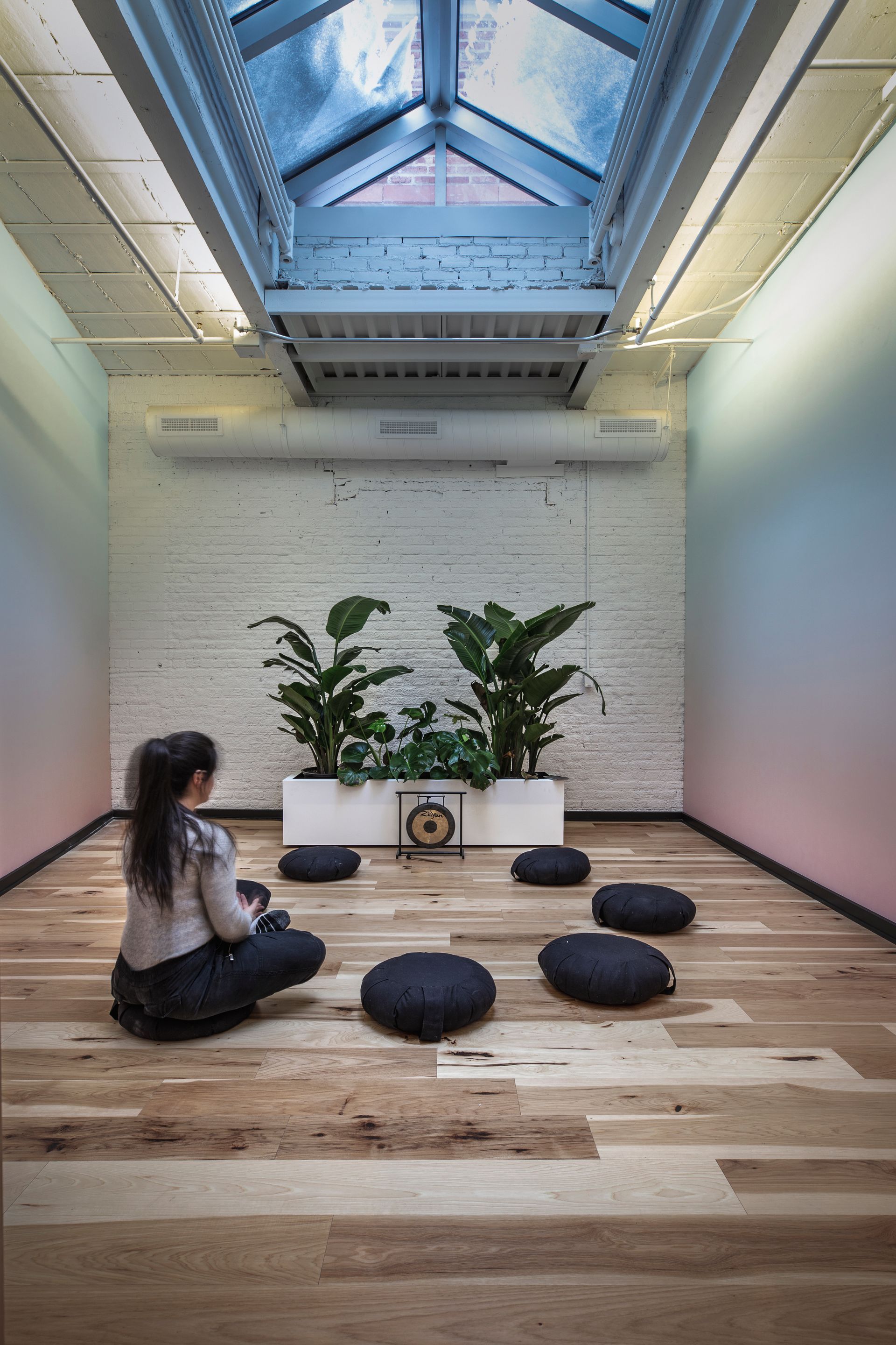 Femme méditant dans une pièce avec des plantes, des coussins et une verrière. Murs aux teintes pastel dégradées.