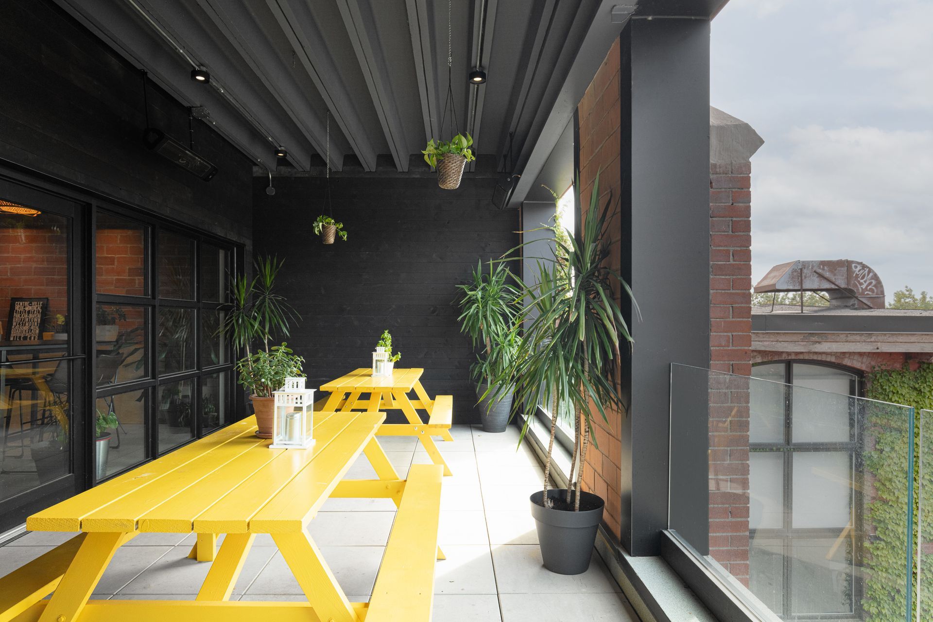 Des tables de pique-nique jaunes sur un balcon moderne avec des plantes et un bâtiment en briques en arrière-plan.