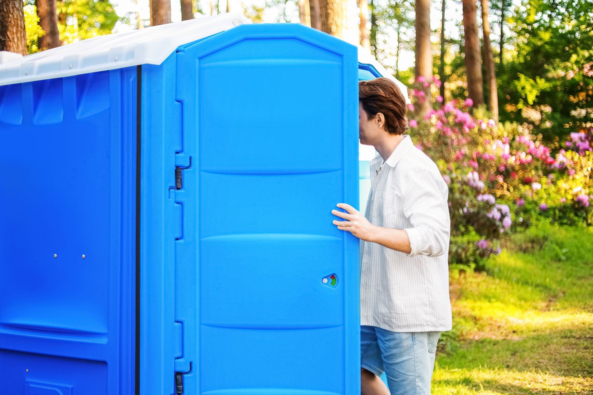 Person opening a bright blue portable restroom outdoors beside flowers and trees