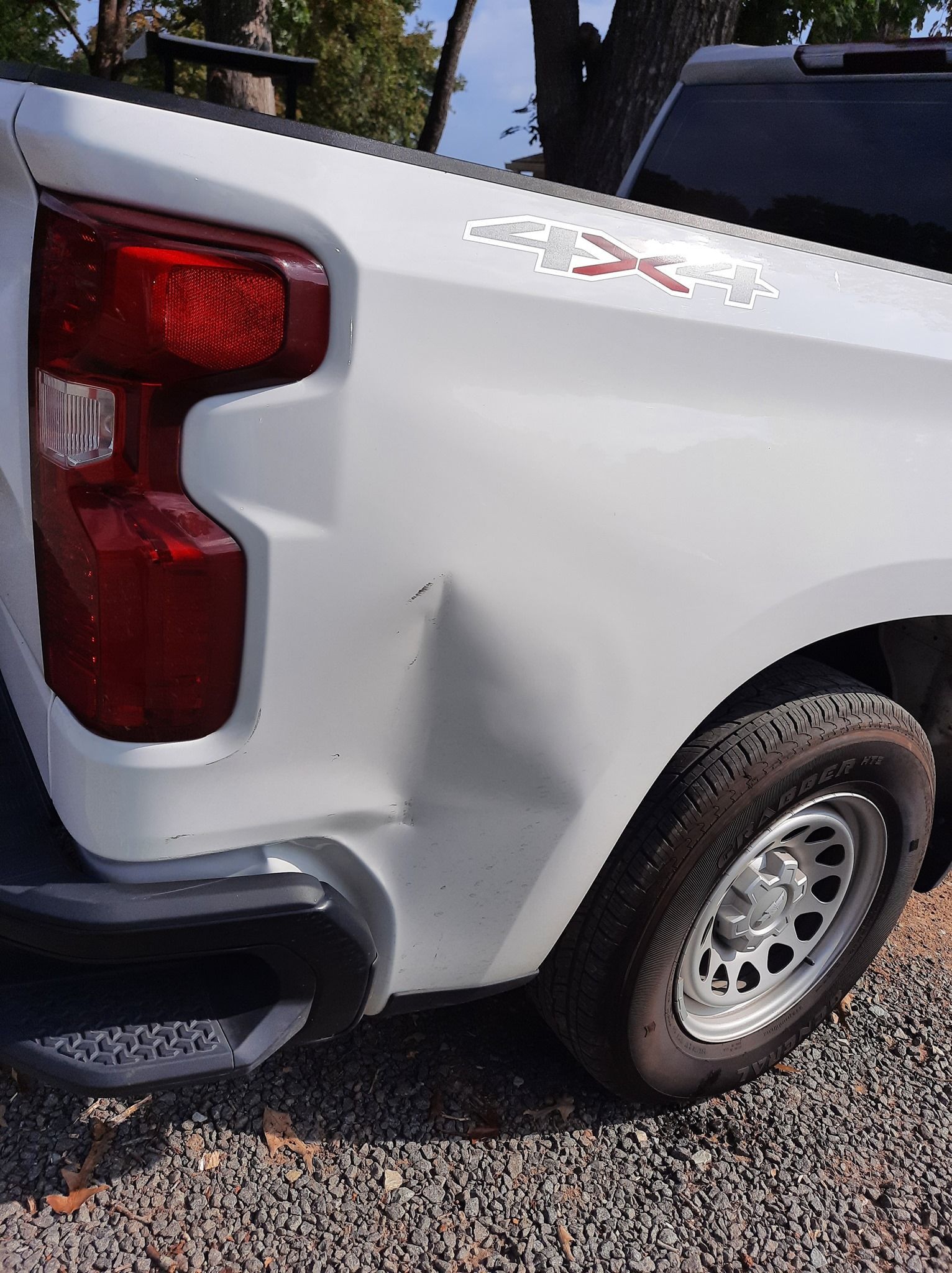 White pickup truck with a dented rear side panel and red taillight parked on gravel