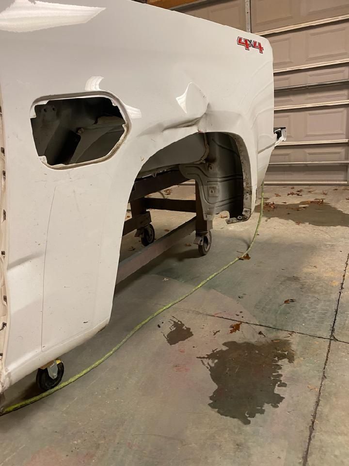 White car body in a garage with a large dented rear quarter panel and exposed wheel well.