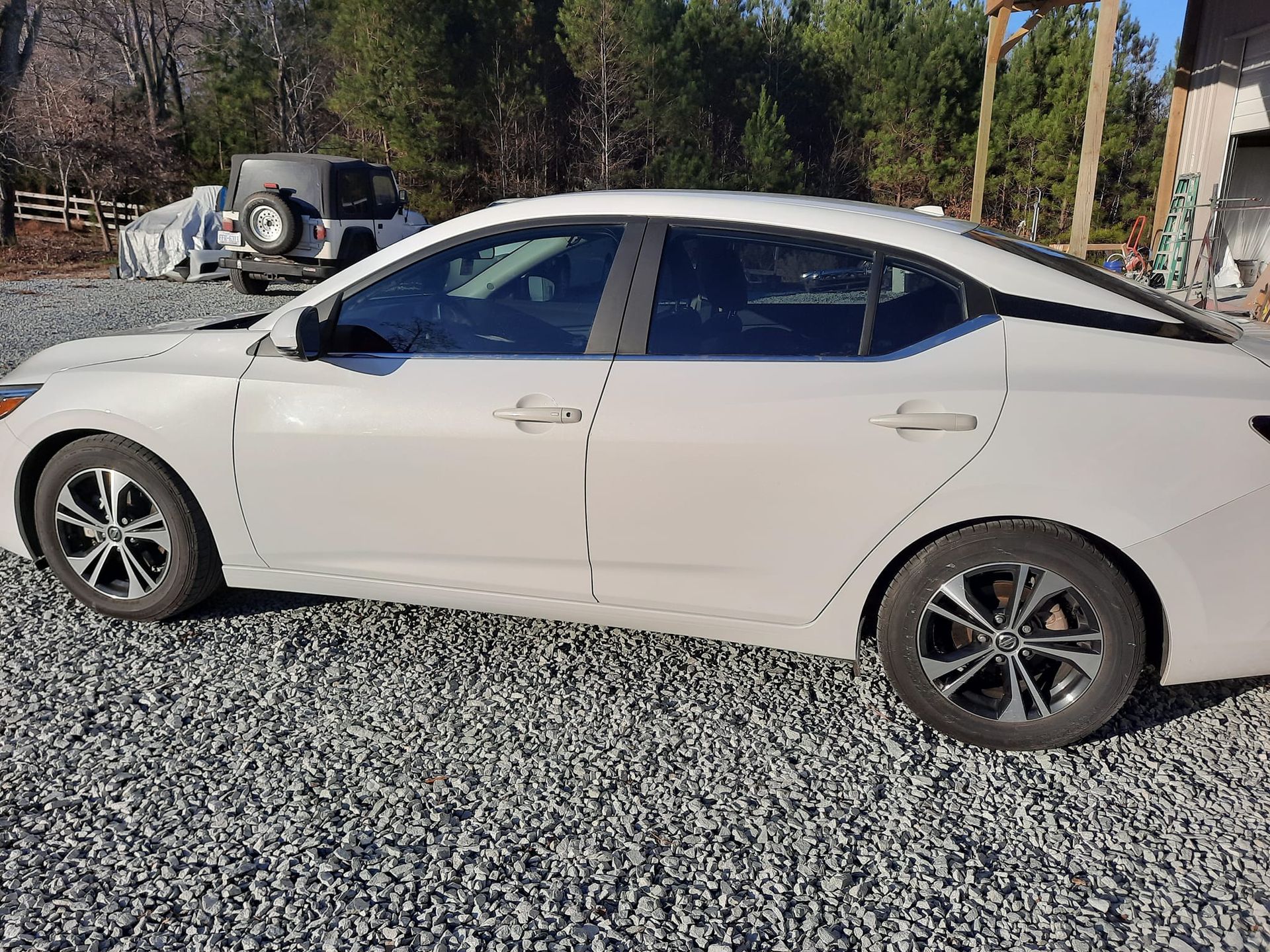 White sedan parked on gravel beside trees in bright daylight
