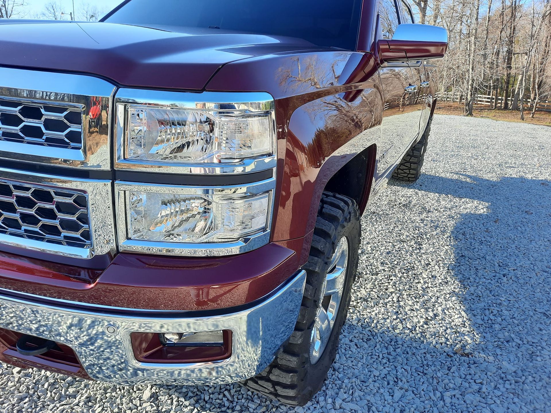 Maroon pickup truck front end parked on gravel, with chrome grille and headlight, in a sunny outdoor lot