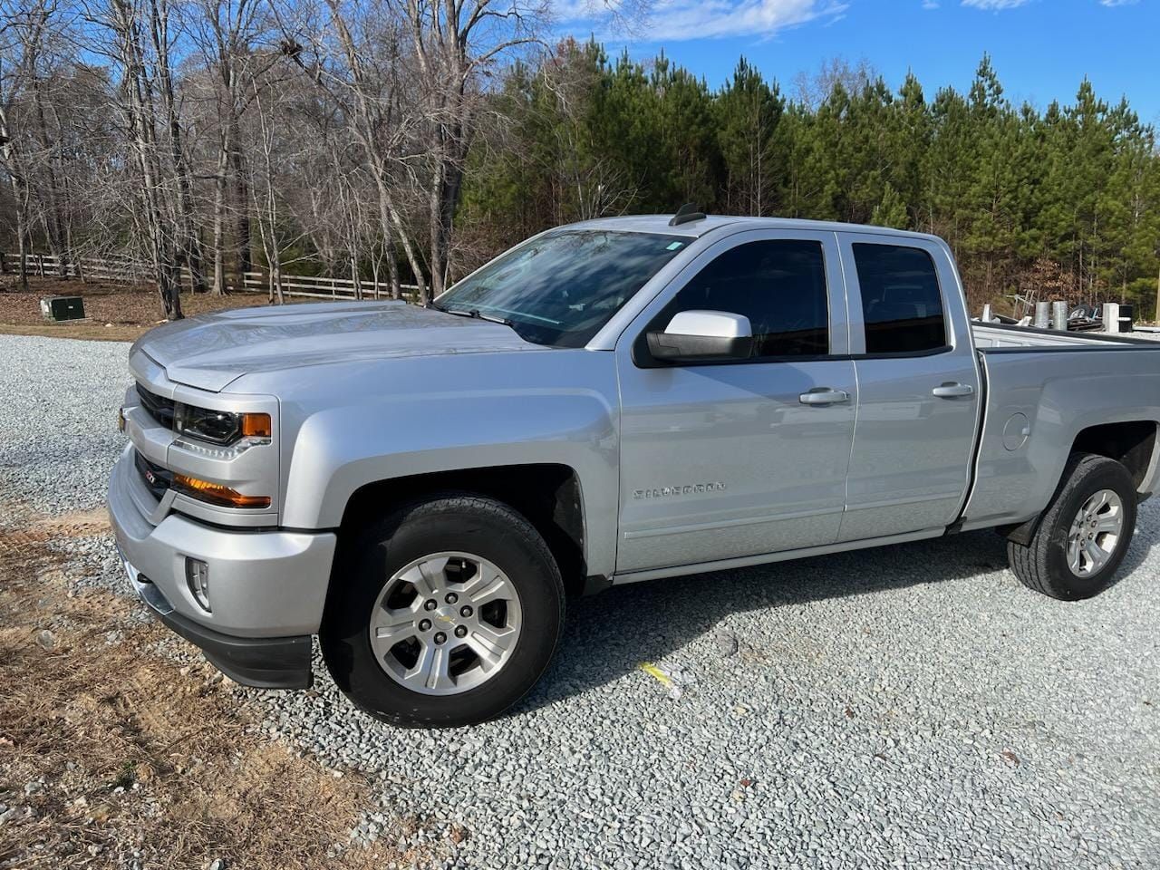 Silver pickup truck parked on a gravel lot beside trees on a sunny day