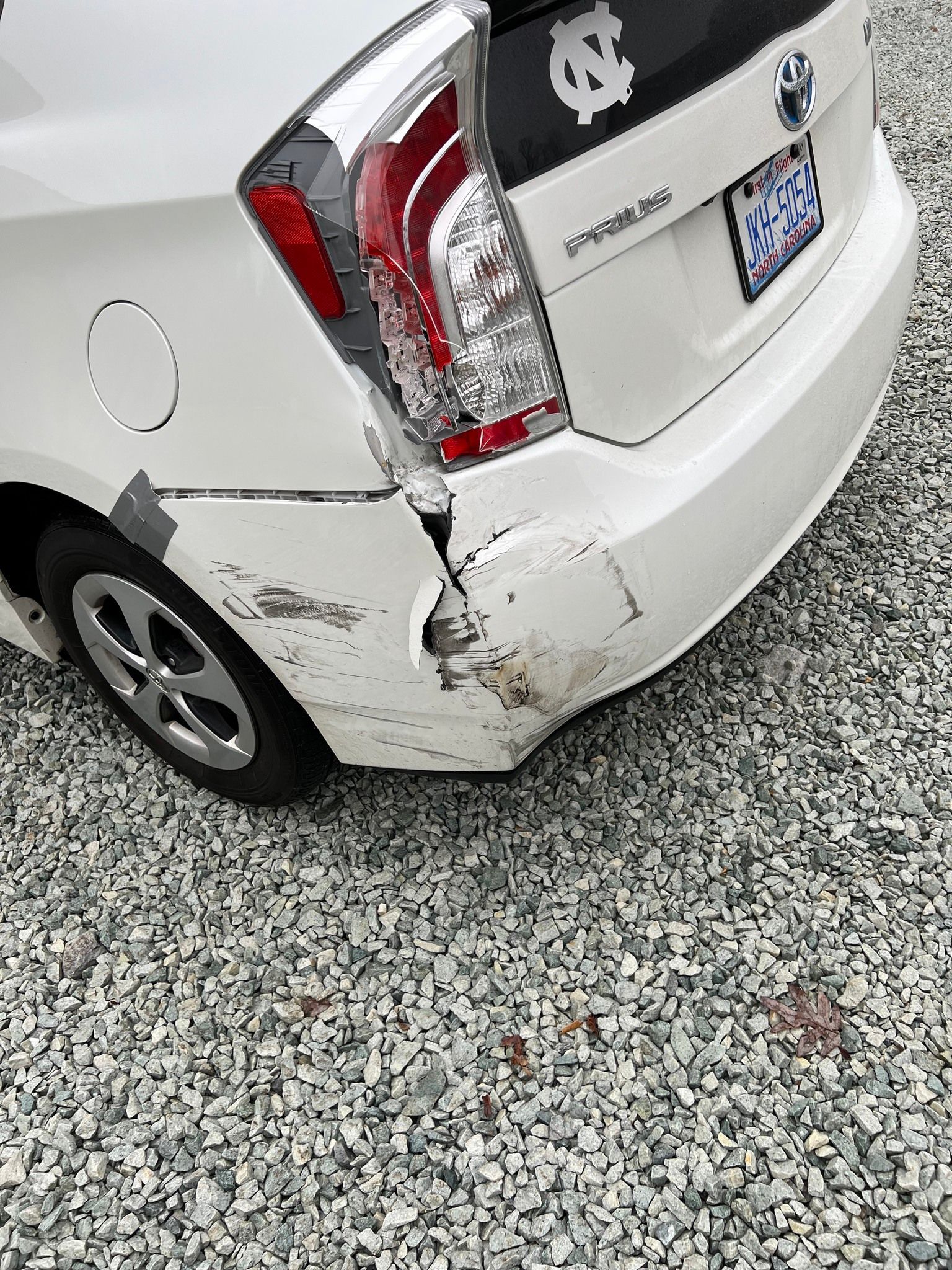 Damaged white car rear bumper and taillight with scraped and bent left corner parked on gravel.