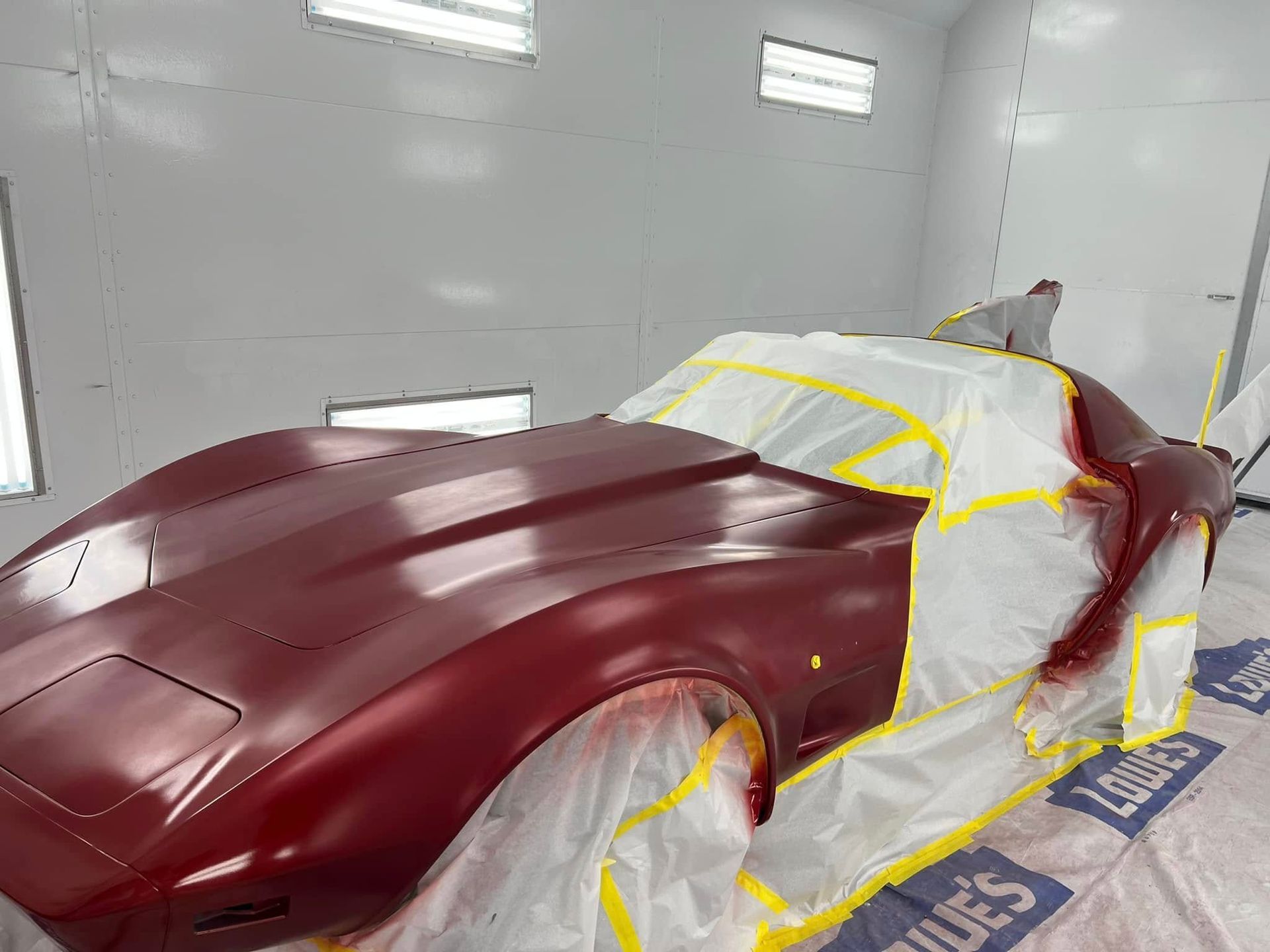 Maroon sports car in a paint booth, masked with tape and plastic for repainting