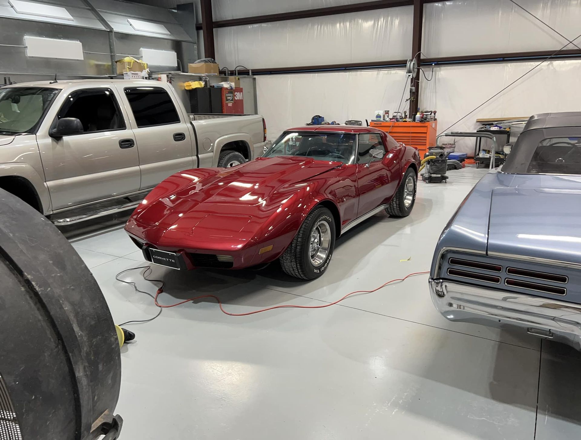 Red classic sports car in a garage, flanked by a silver truck and a gray car.