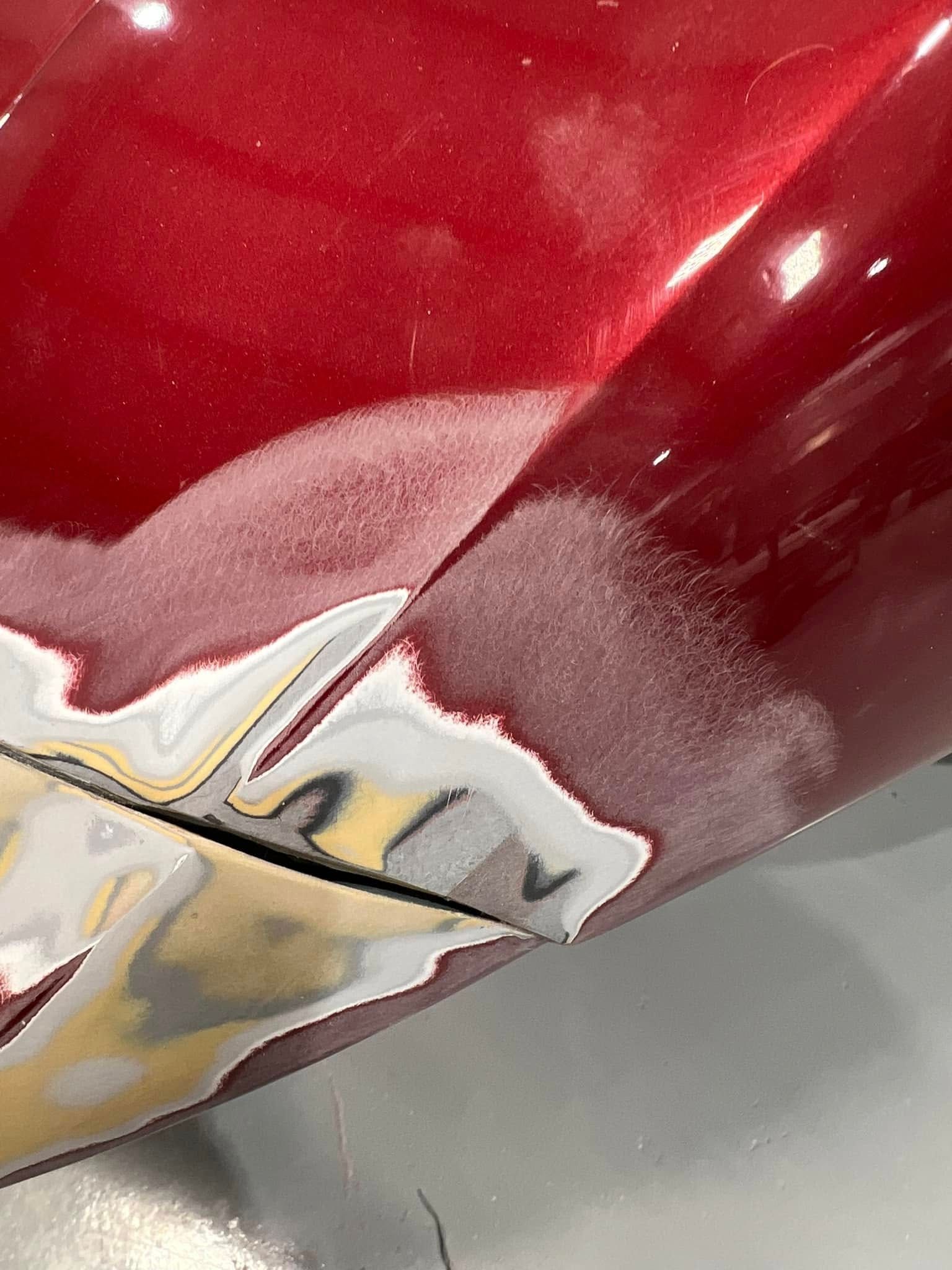 Close-up of a glossy red car body with a chrome emblem and visible paint damage near the panel seam.