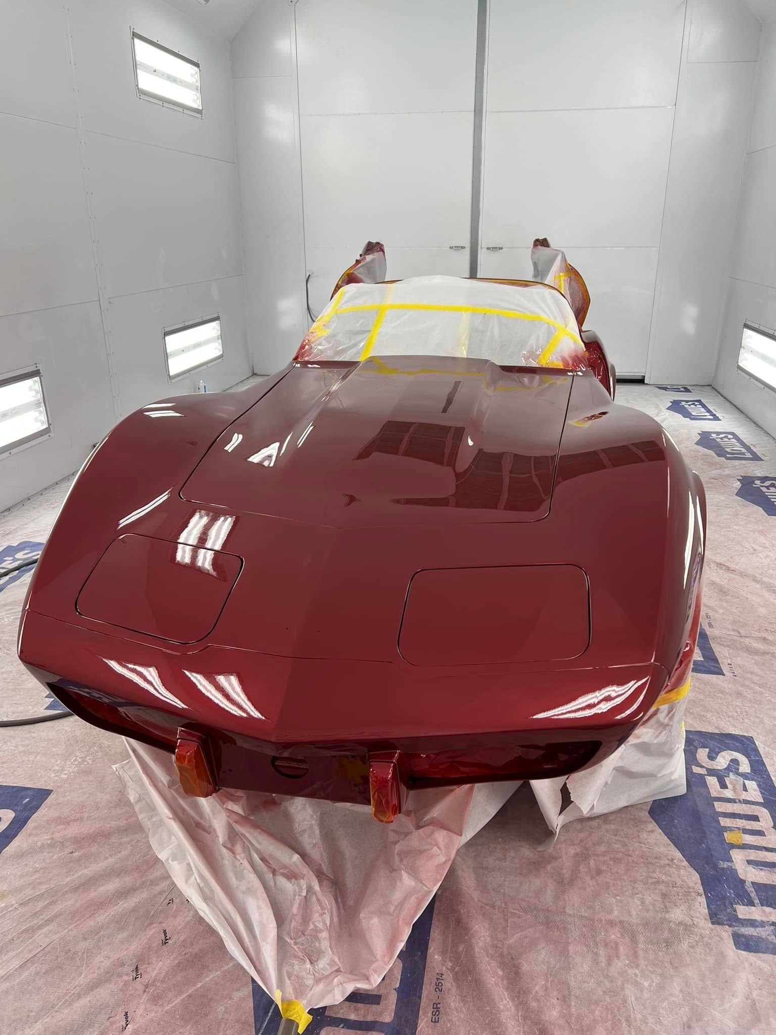 Red classic convertible in a white auto spray booth, partially covered with protective masking.