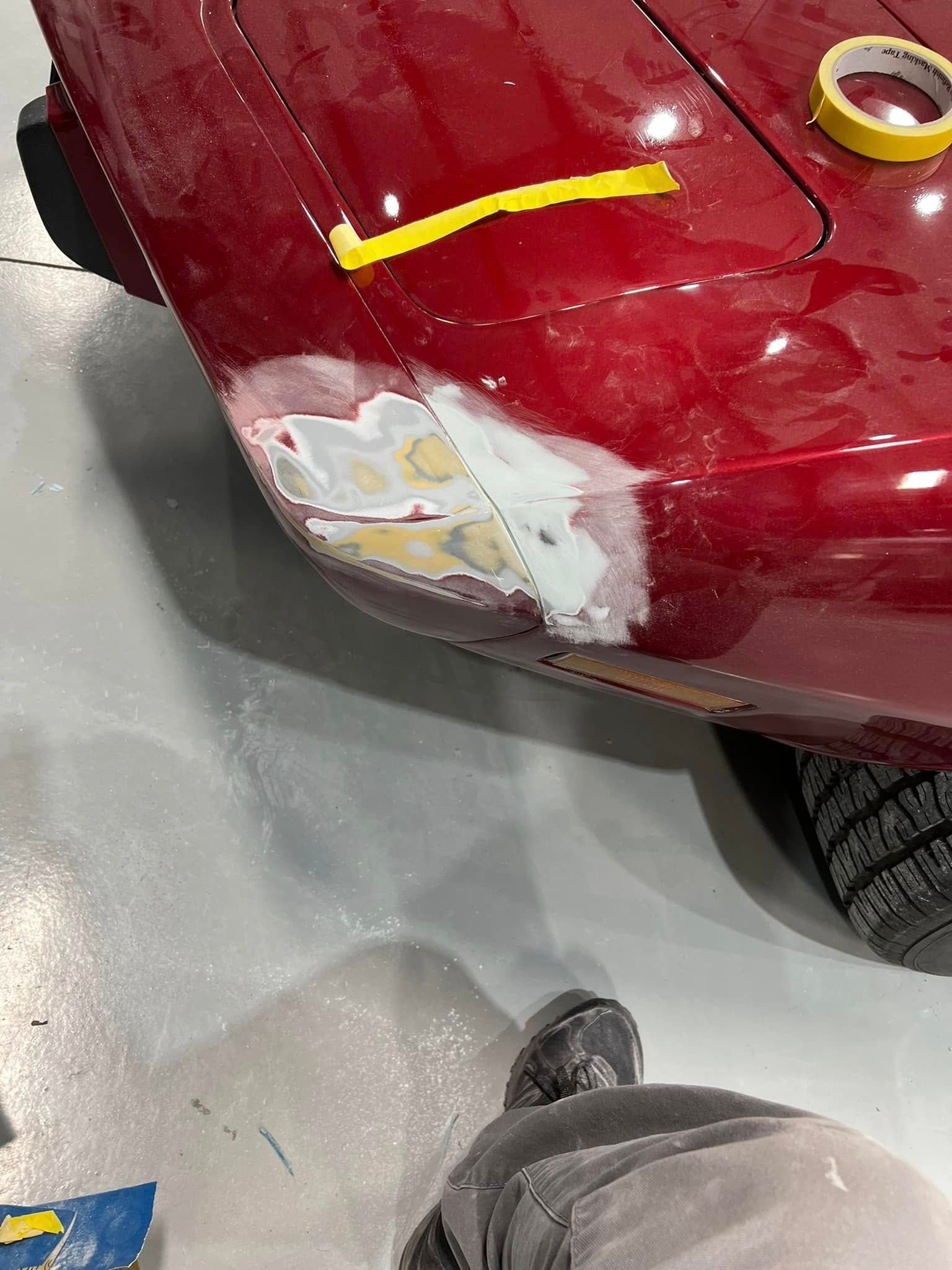 Red car bumper with scraped paint and a patch of body filler near the front wheel