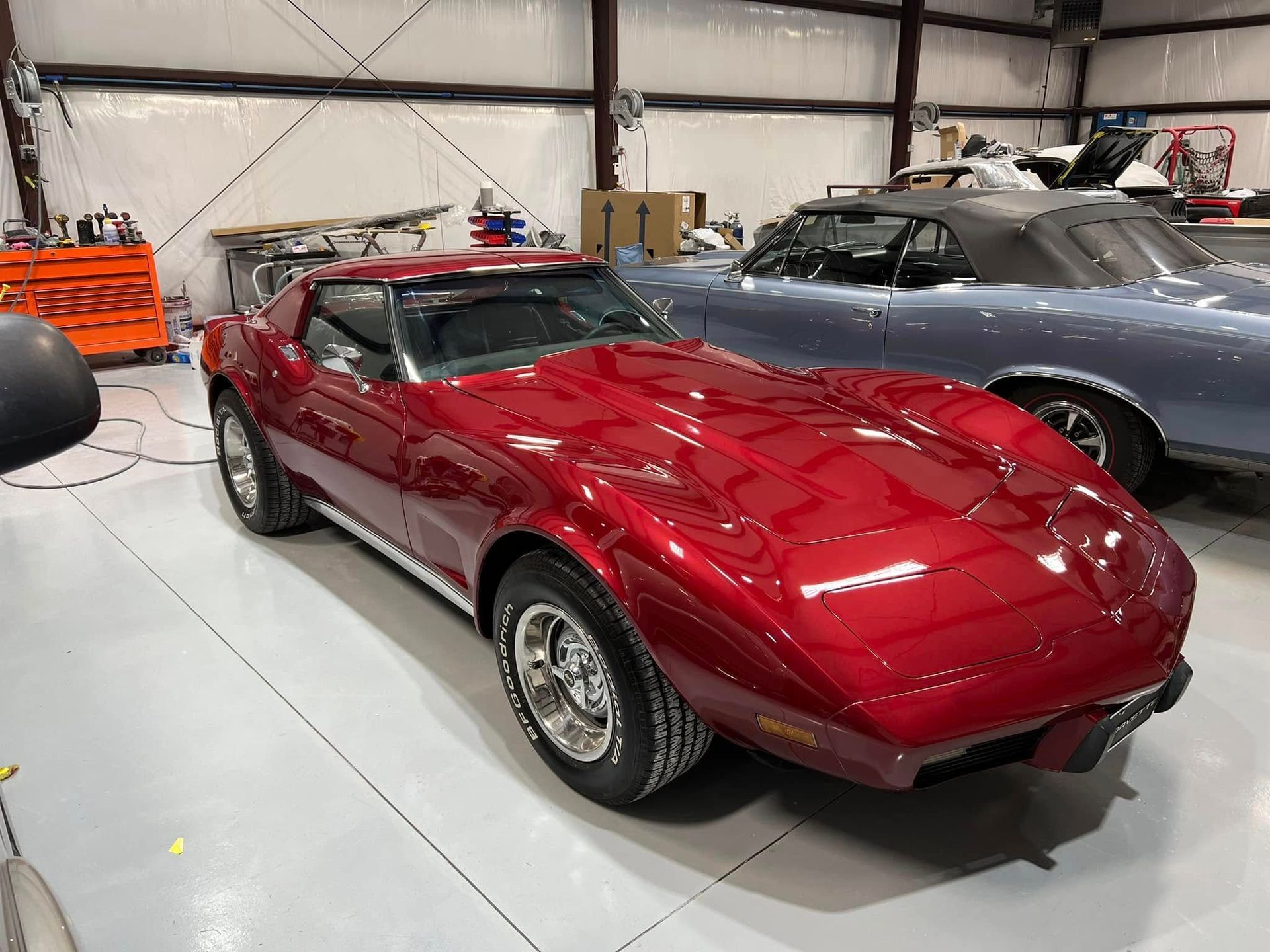 Red classic sports car in a garage, with a gray car parked beside it.