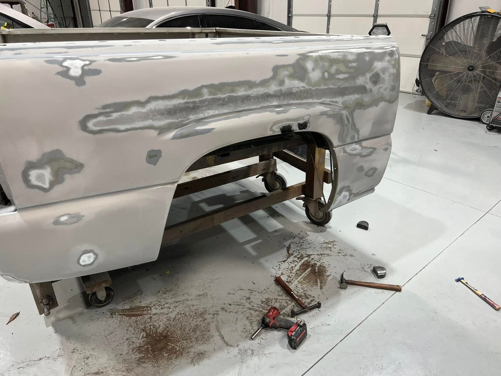 Car body panel in primer with sanding patches, tools, and debris on a workshop floor