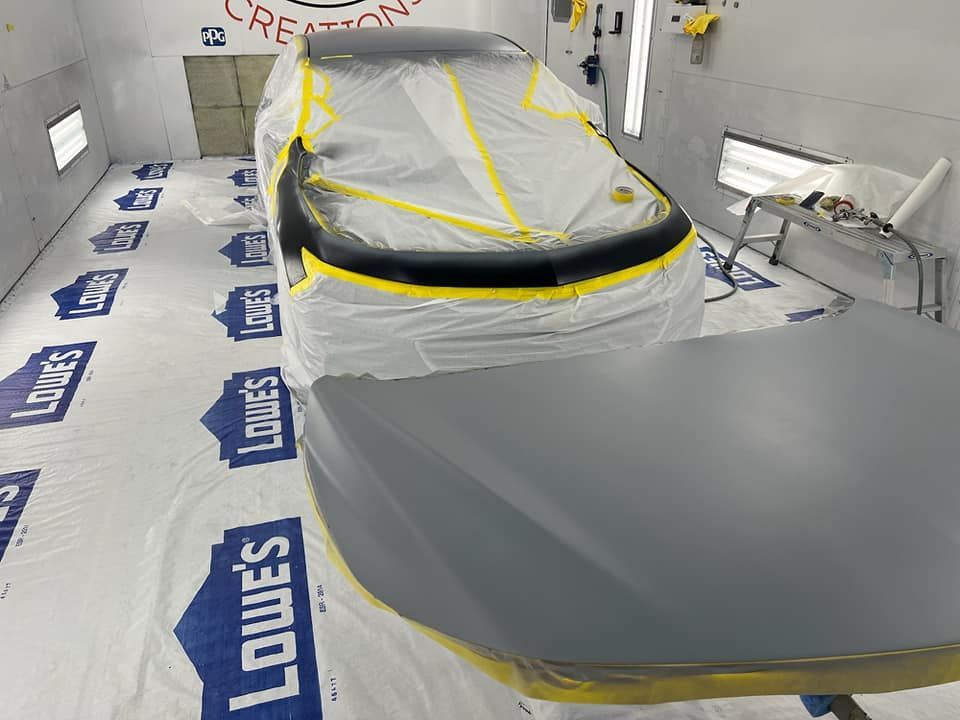 Covered car in auto body shop, masked with plastic and tape beside a painted hood on the floor