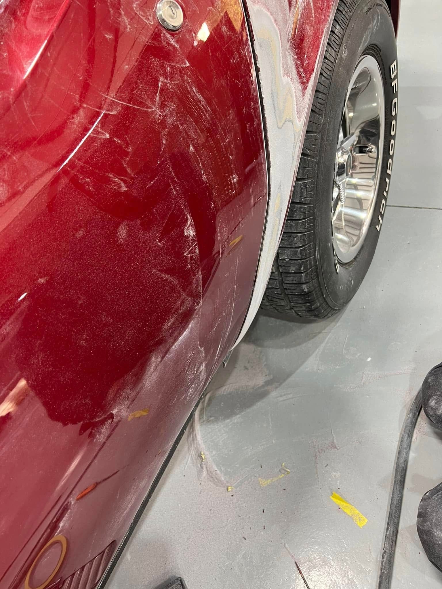 Close-up of a red car’s side with scratches and a rear wheel visible on a garage floor.
