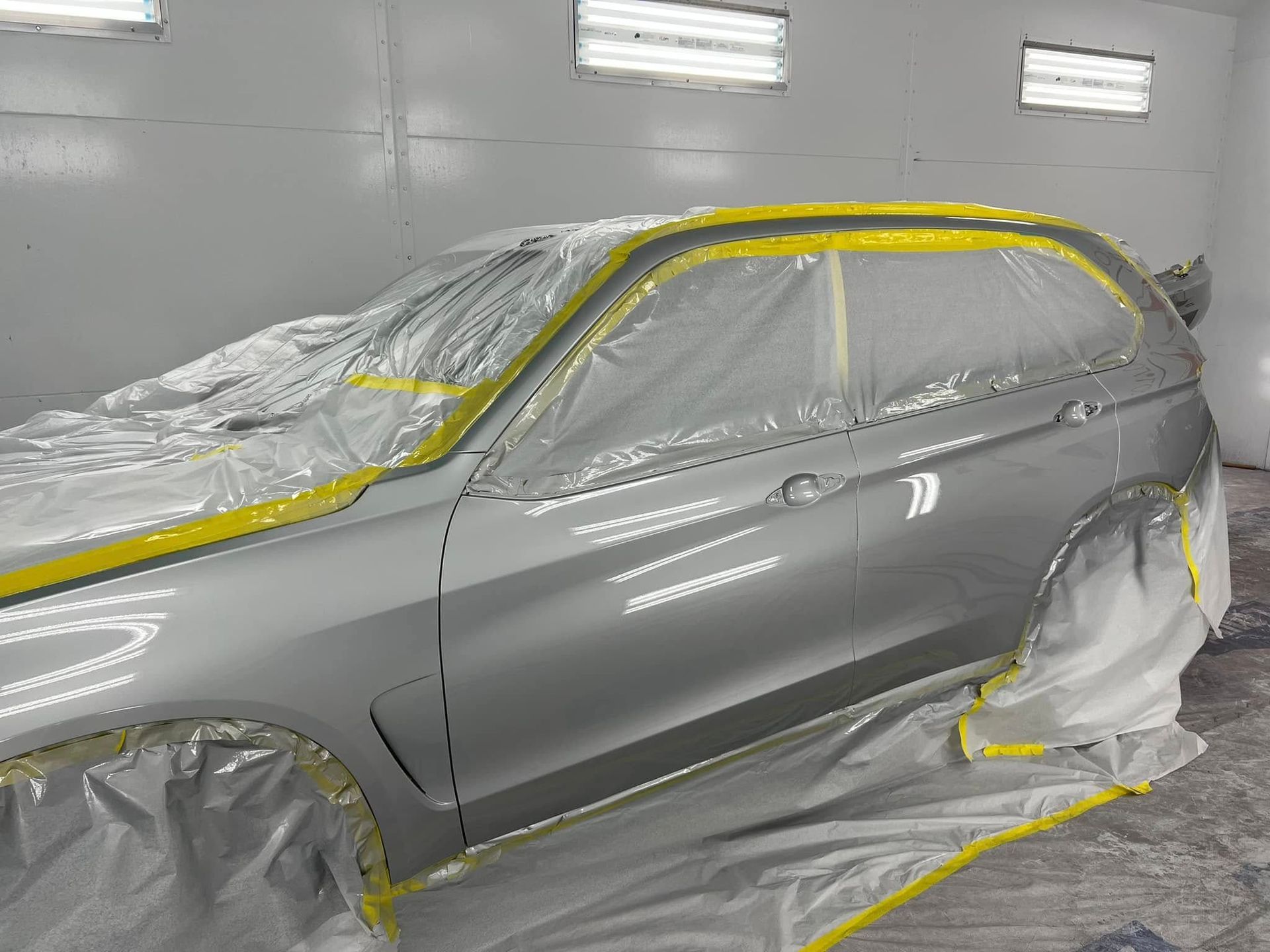 Gray SUV masked for painting in a spray booth, with yellow tape along the windows and trim.
