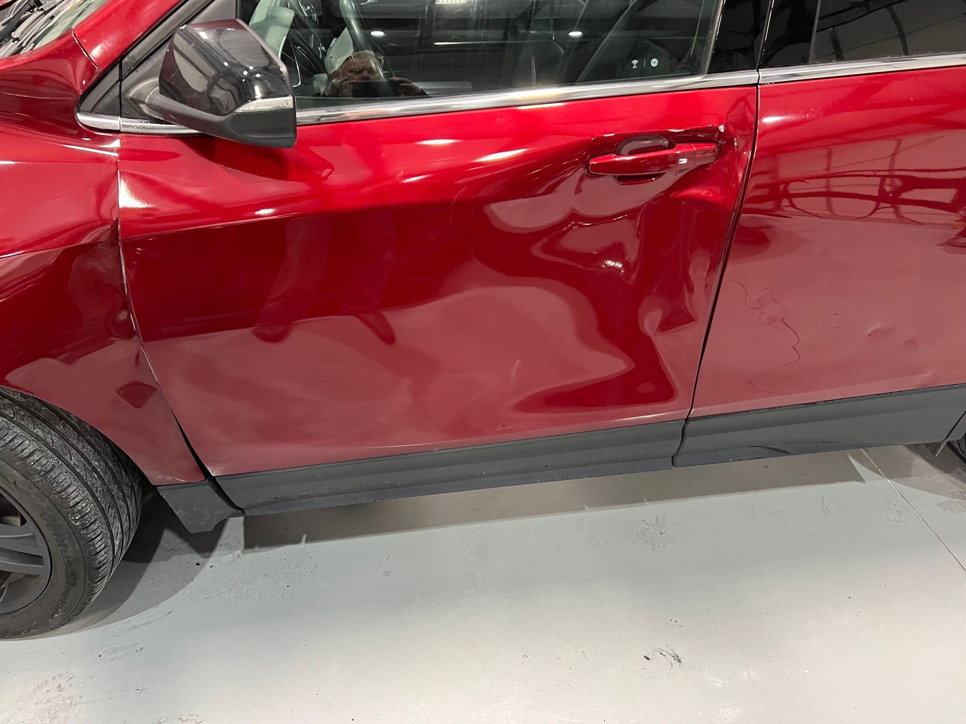 Red SUV door with a large dent and scrape near the handle in a garage or shop.