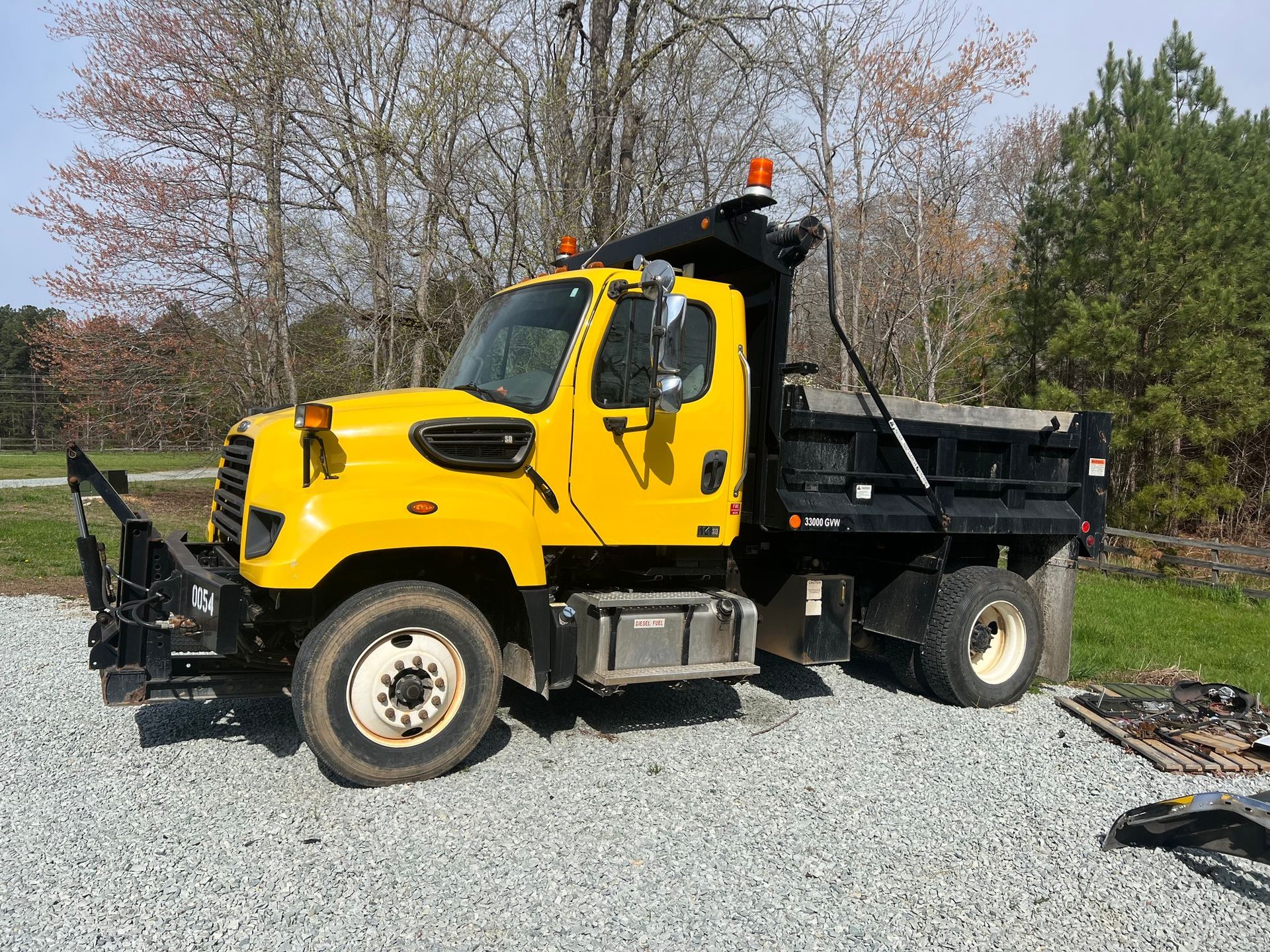 Yellow dump truck parked on gravel in a wooded lot under a clear sky