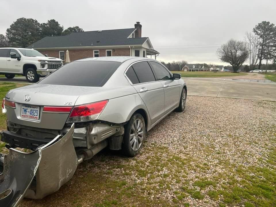 Silver sedan with rear-end damage, bumper detached, parked on a gravel lot near a house.