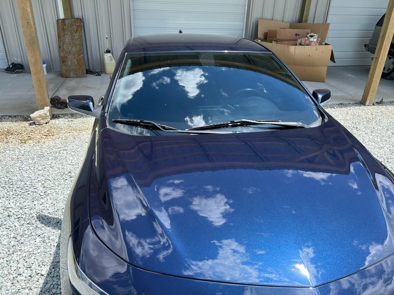 Blue car parked in front of a garage, with reflections on the hood and windshield.