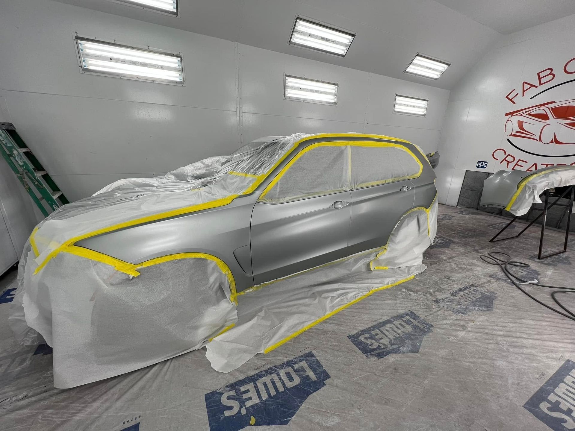 Gray car body in a paint booth, covered with masking plastic and outlined in yellow tape.