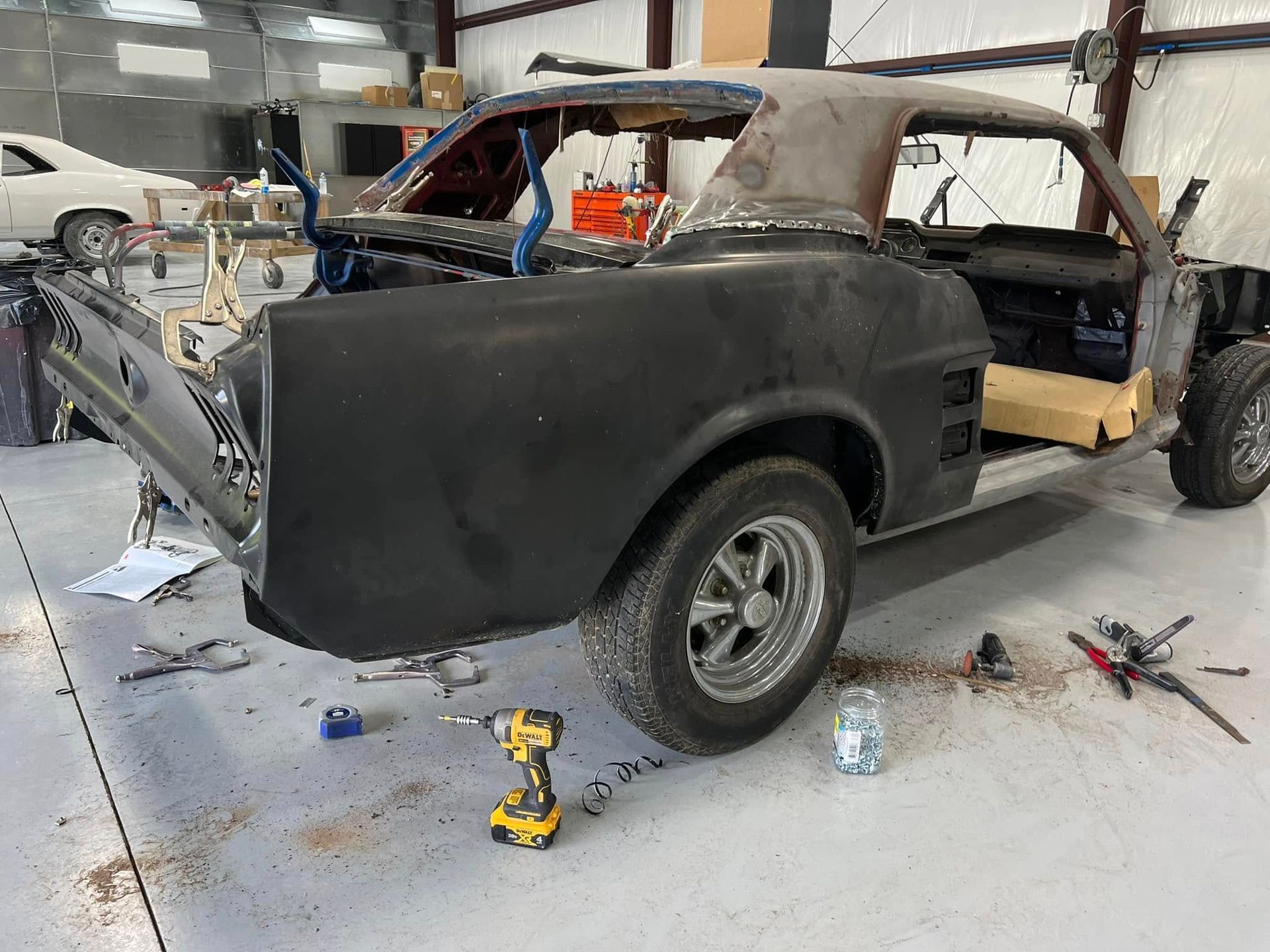 Vintage coupe under restoration in a garage, with black primer body, exposed roof frame, and tools on the floor.